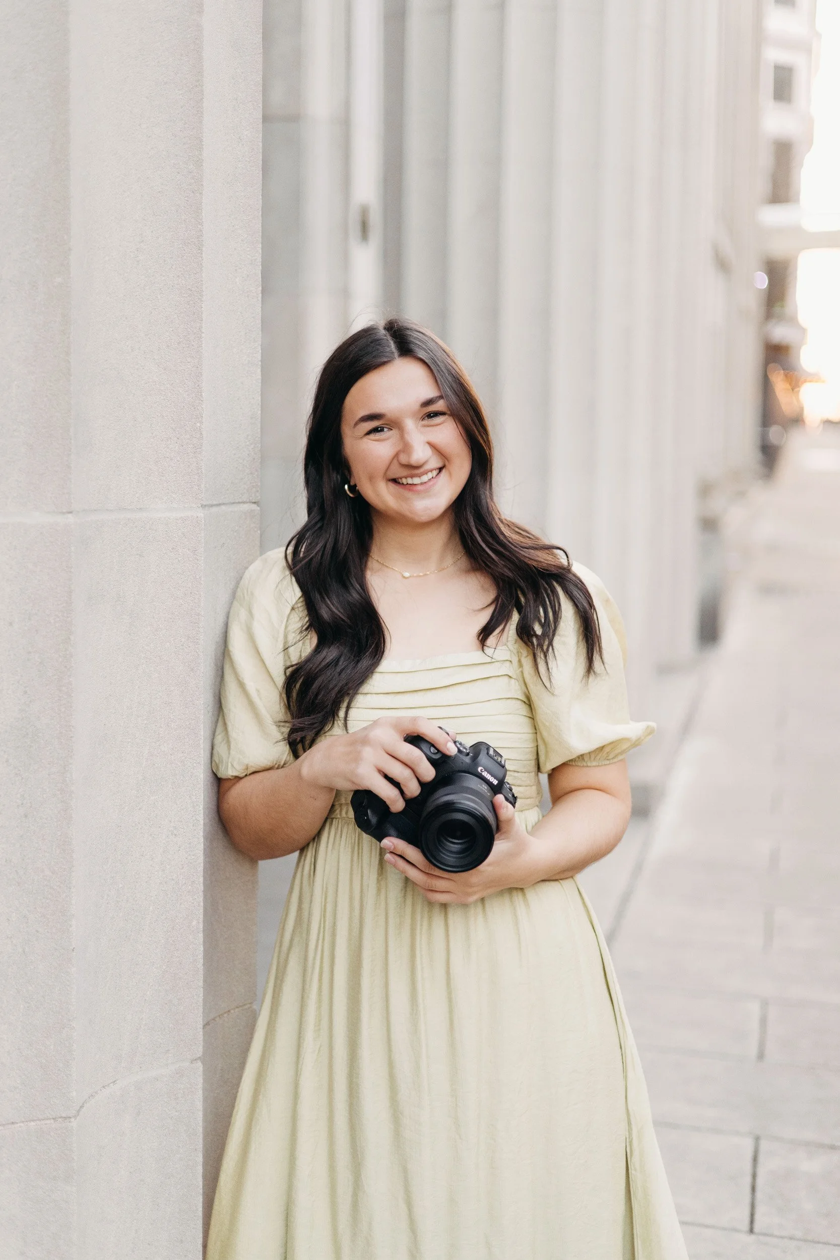 A smiling woman with long dark hair holding a professional camera in her hands, standing outdoors against a city street with light-colored building walls.