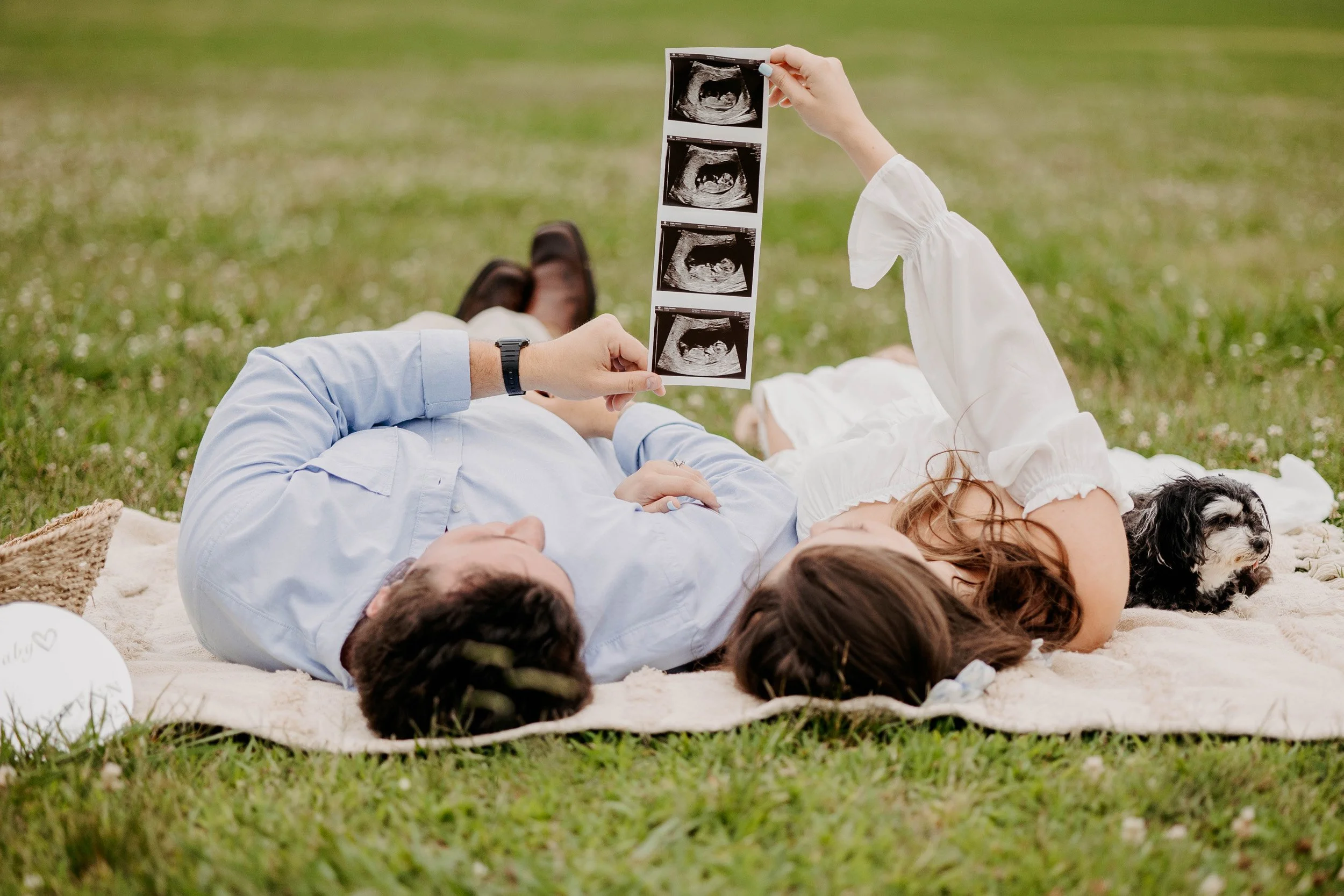 A couple lying on a blanket outdoors on grass, holding up ultrasound photos of a baby, with a small dog nearby.