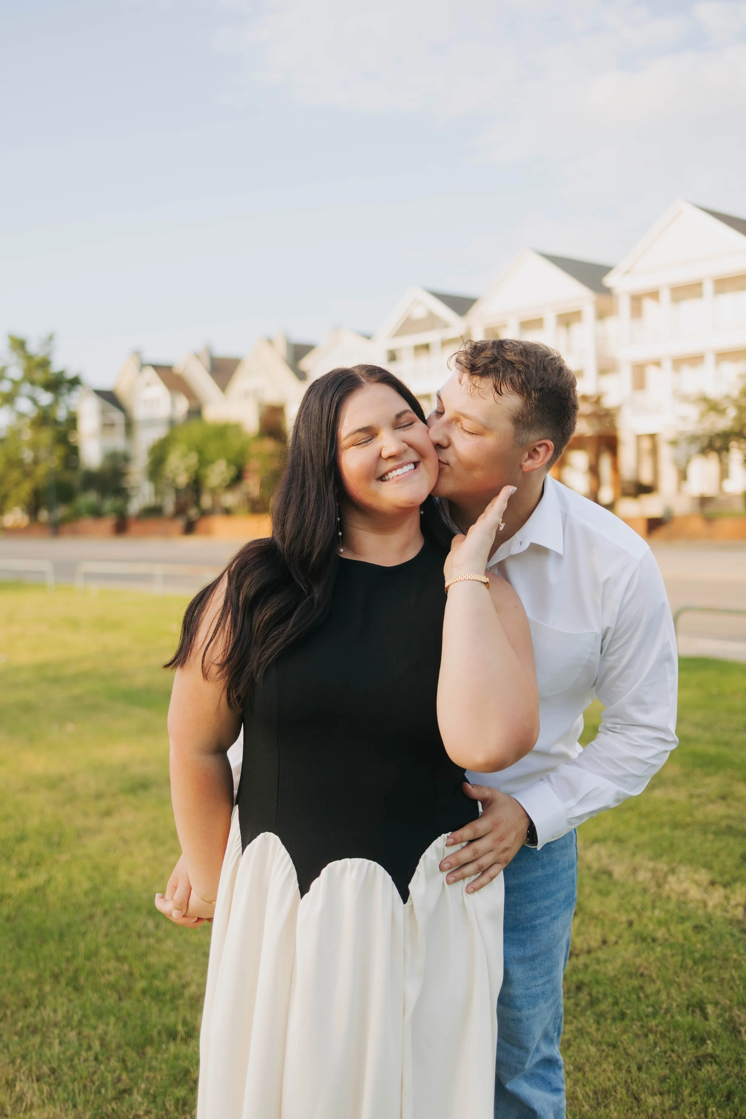 A happy couple outside on a grassy area with a row of white houses in the background. The man is kissing the woman on her cheek while she smiles with her eyes closed, wearing a black and white dress. The man is dressed in a white shirt and jeans, hol