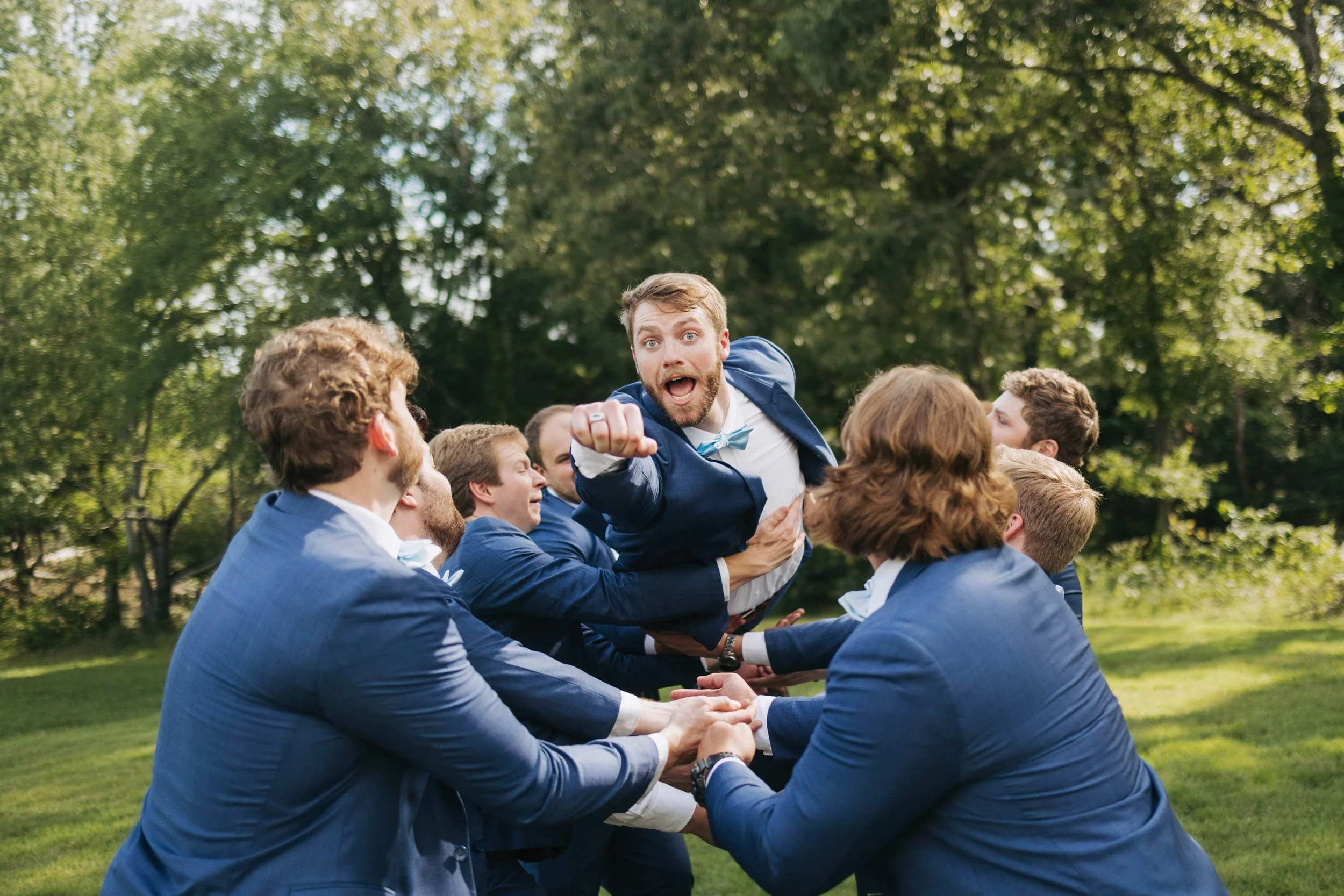 A group of smiling men in suits forming a human circle outdoors, with one man in a blue suit and bow tie being lifted by others.