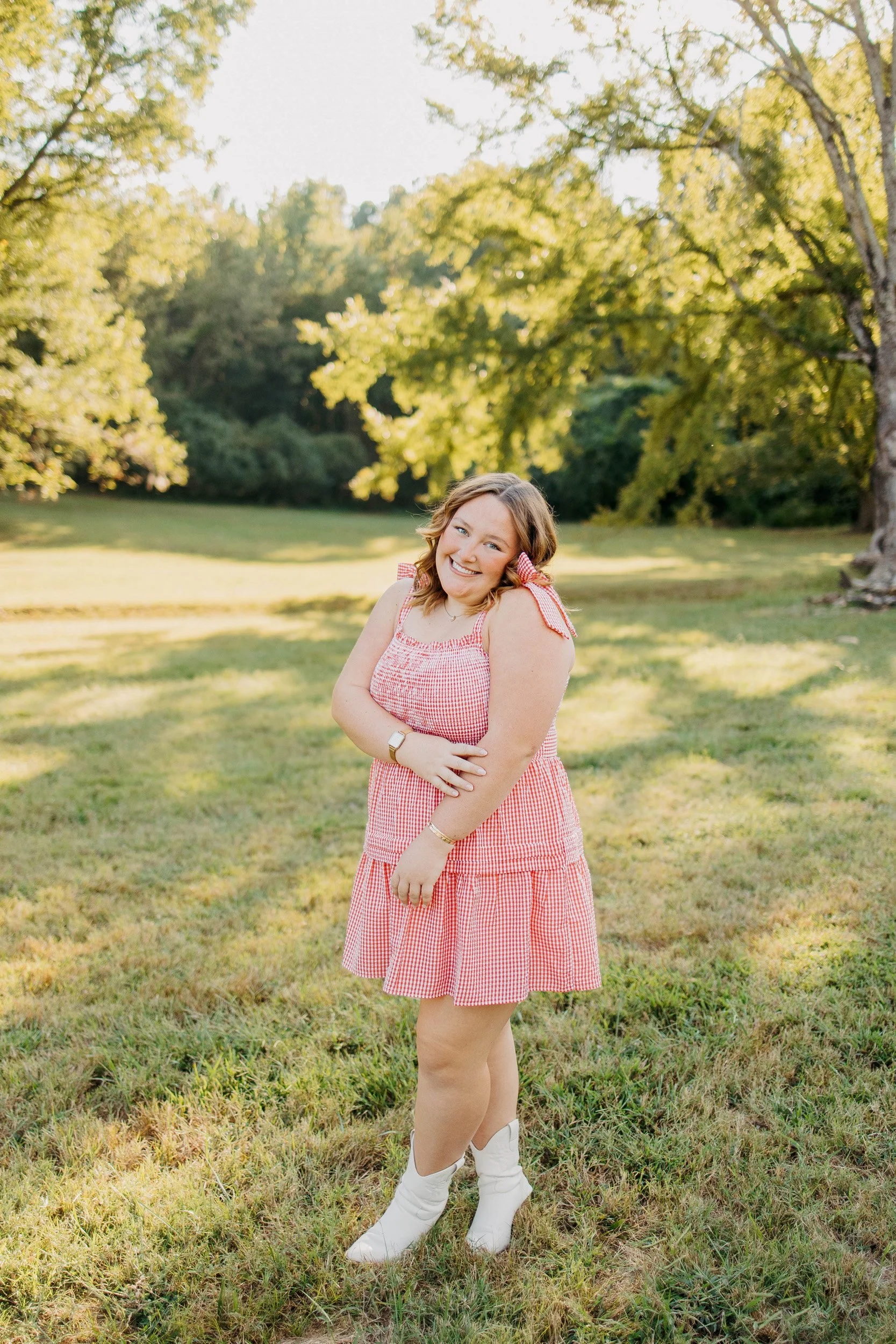 Young woman with shoulder-length hair smiling and standing outdoors in a grassy park with trees in the background, wearing a red gingham dress and white cowboy boots.