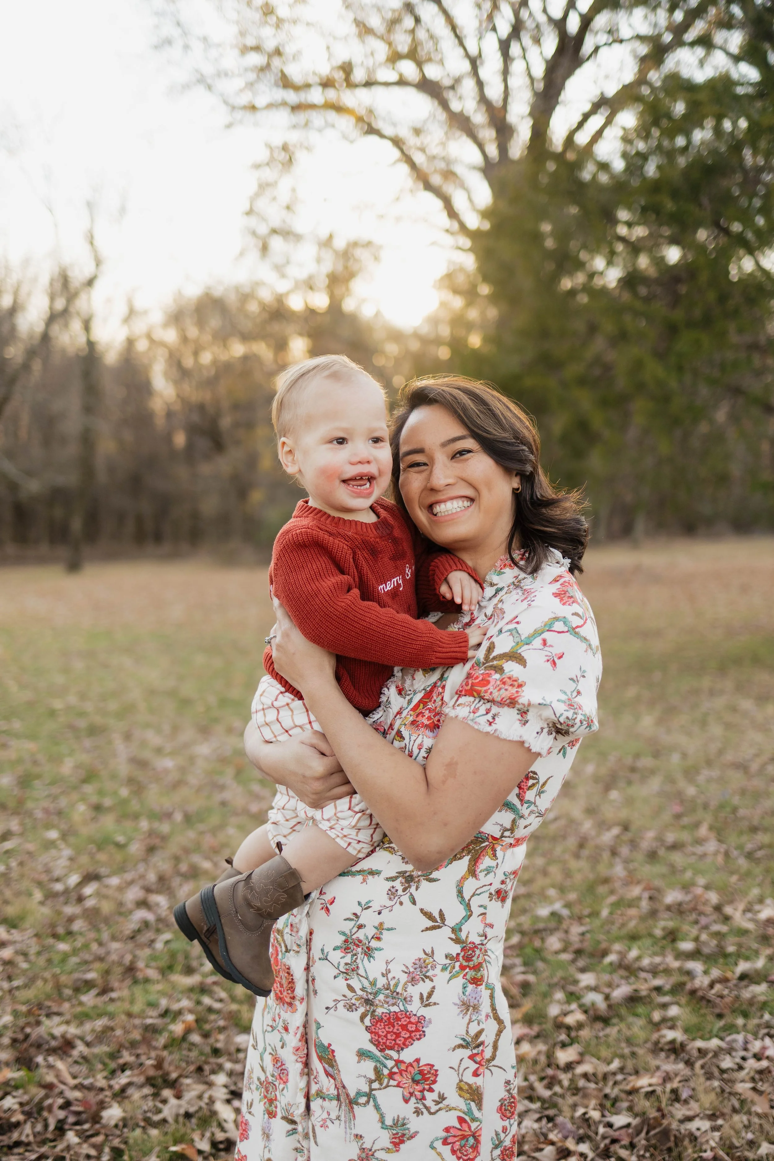A woman holding a smiling young boy outdoors in a park during autumn, with trees and fallen leaves in the background, during sunset.