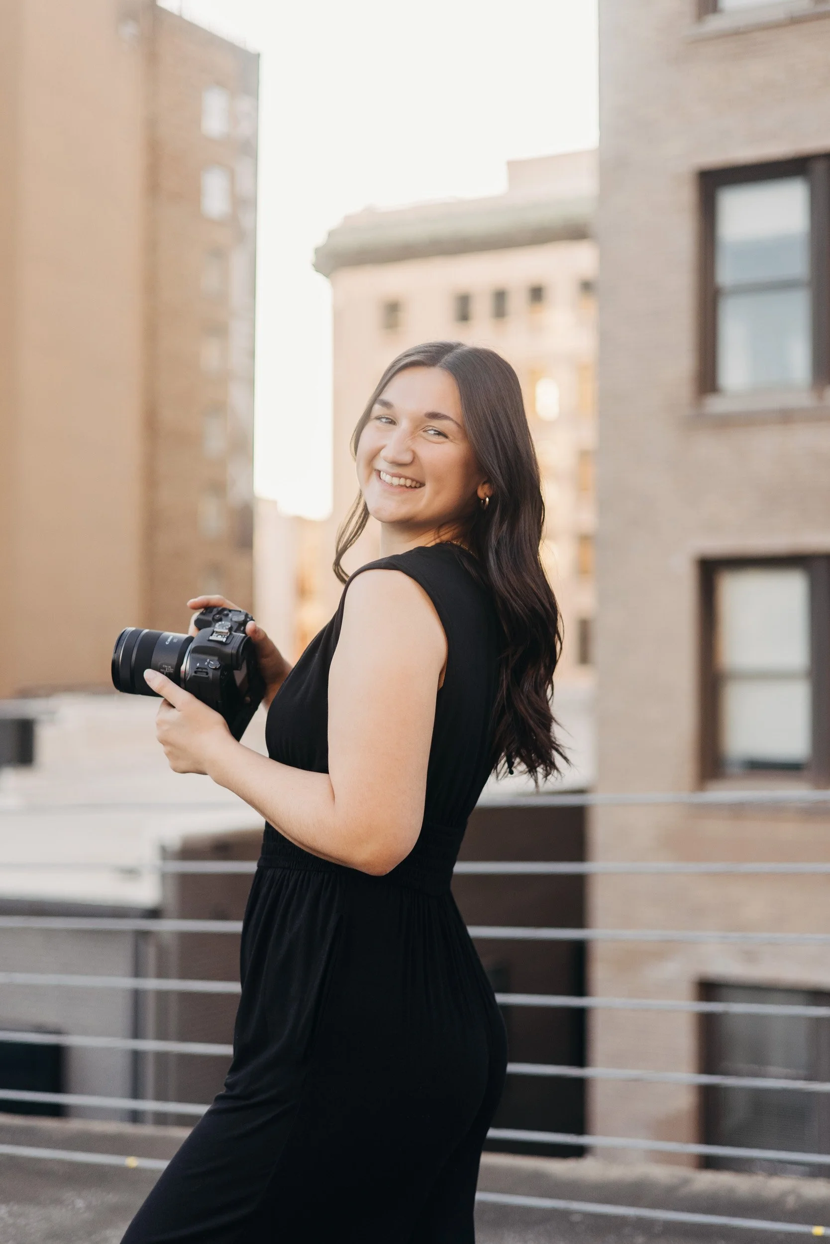 A woman with long dark hair smiling and holding a camera, wearing a black dress, standing outdoors in an urban setting.