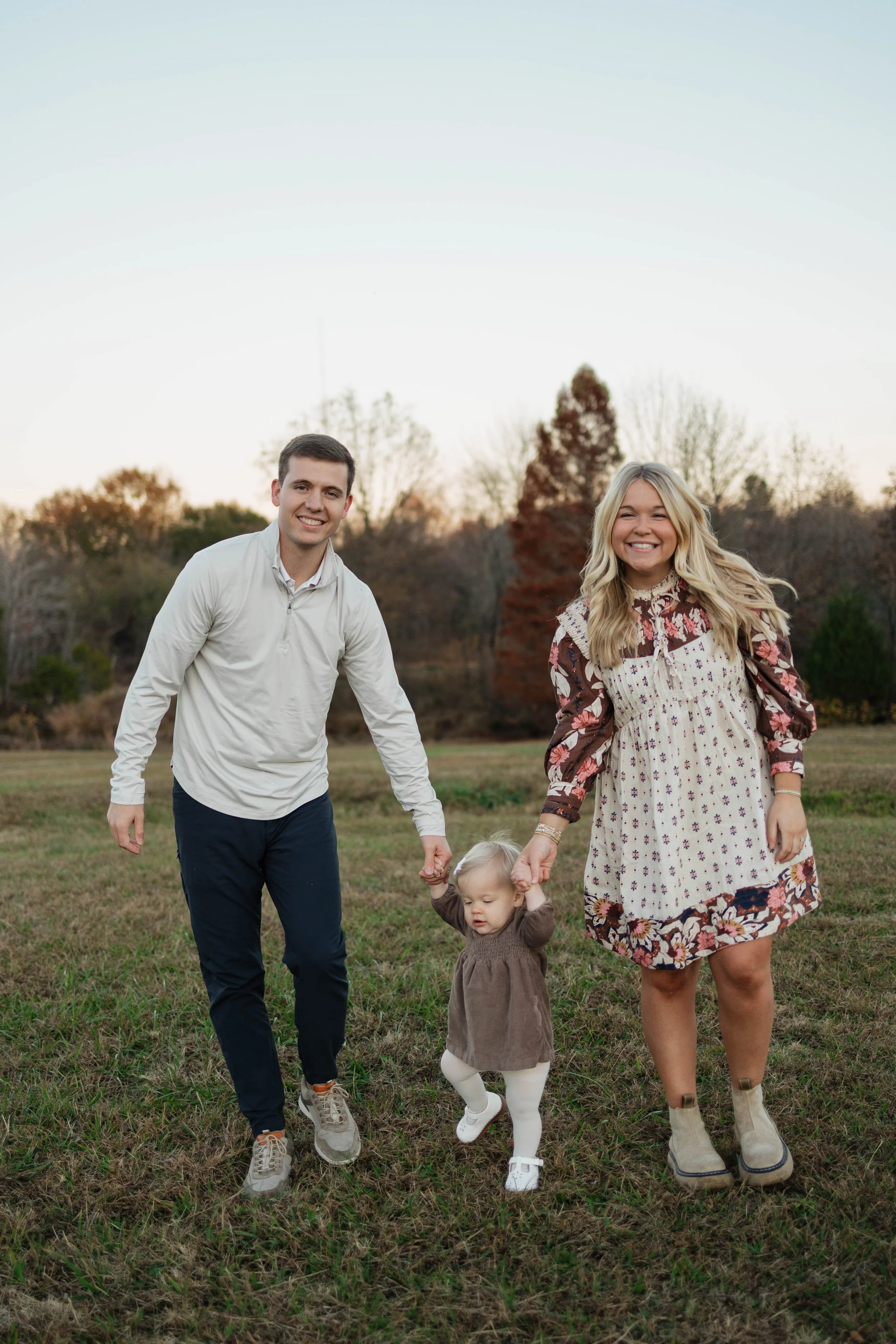 A family of four walking in a grassy park during sunset, with trees in the background. The father is holding the hand of a young girl in a brown dress, while the mother holds her other hand. The father has short dark hair and is wearing a white zip-u