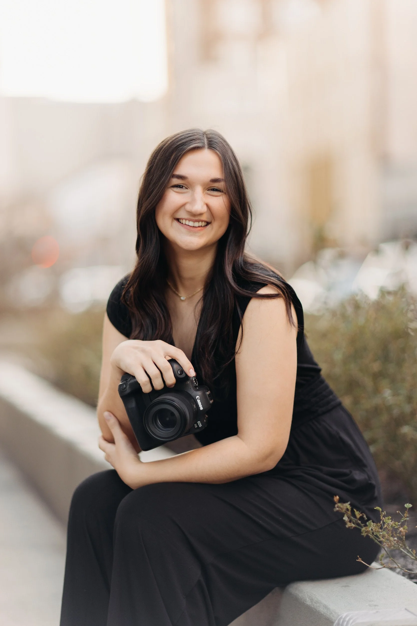 A smiling young woman with long, dark, wavy hair sitting outdoors and holding a Canon camera in her hands.