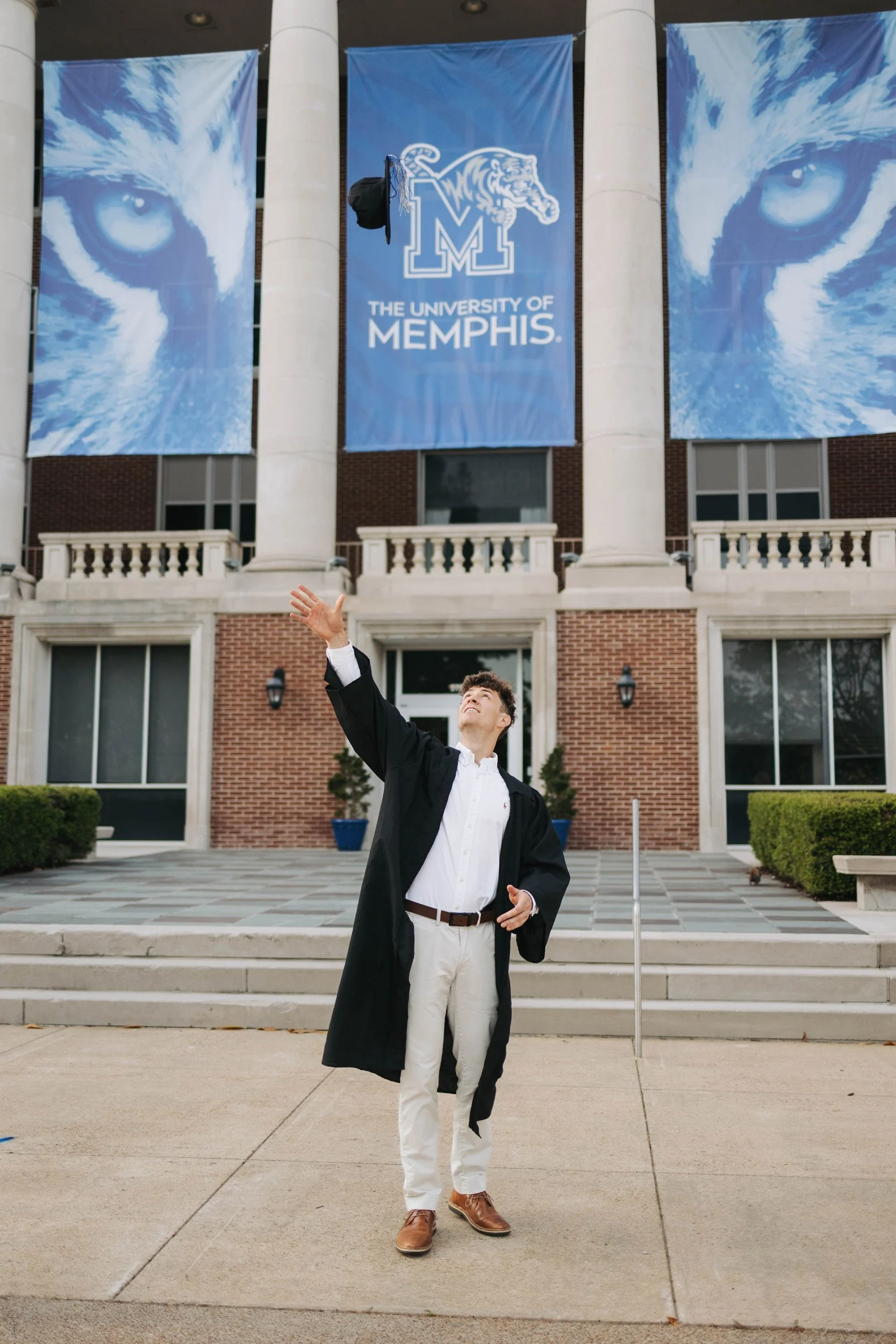 A young man in graduation attire standing in front of the University of Memphis building, waving and smiling. The building has large banners featuring a close-up of a tiger's face and the university's logo.