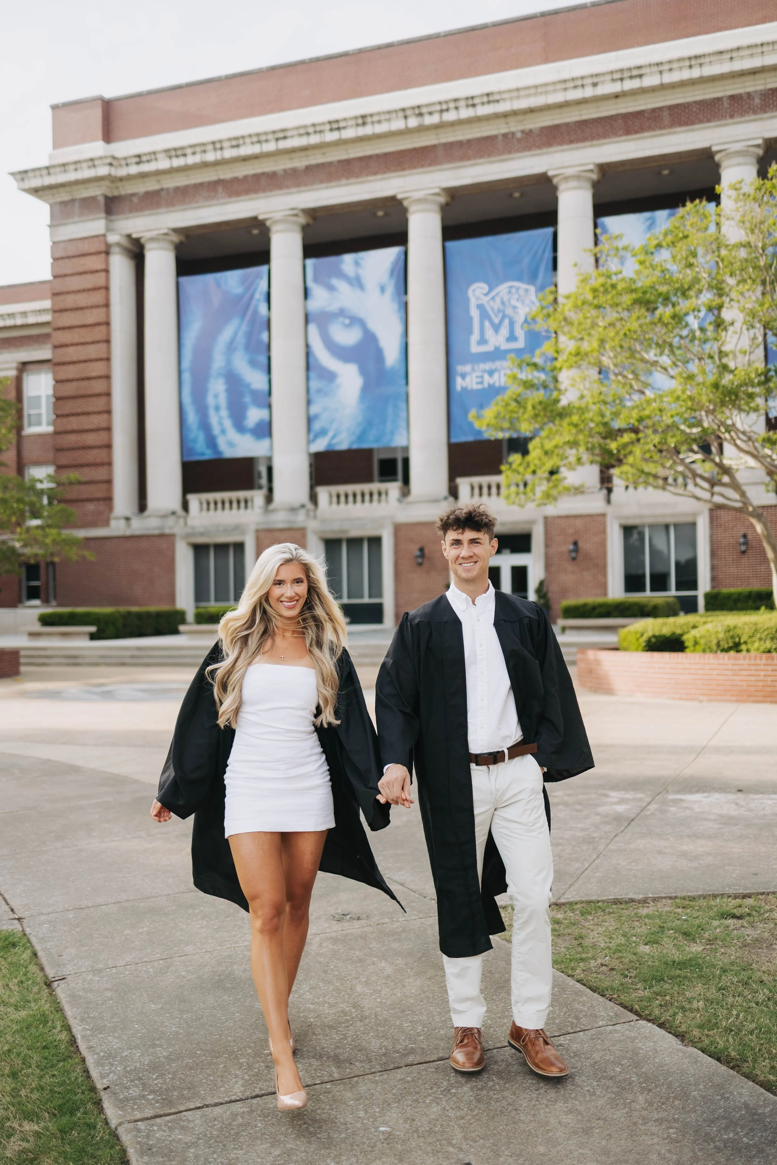 A young woman and man in graduation gowns walking on a sidewalk in front of a brick building with columns. The building has blue banners with a tiger mascot and text related to a university.