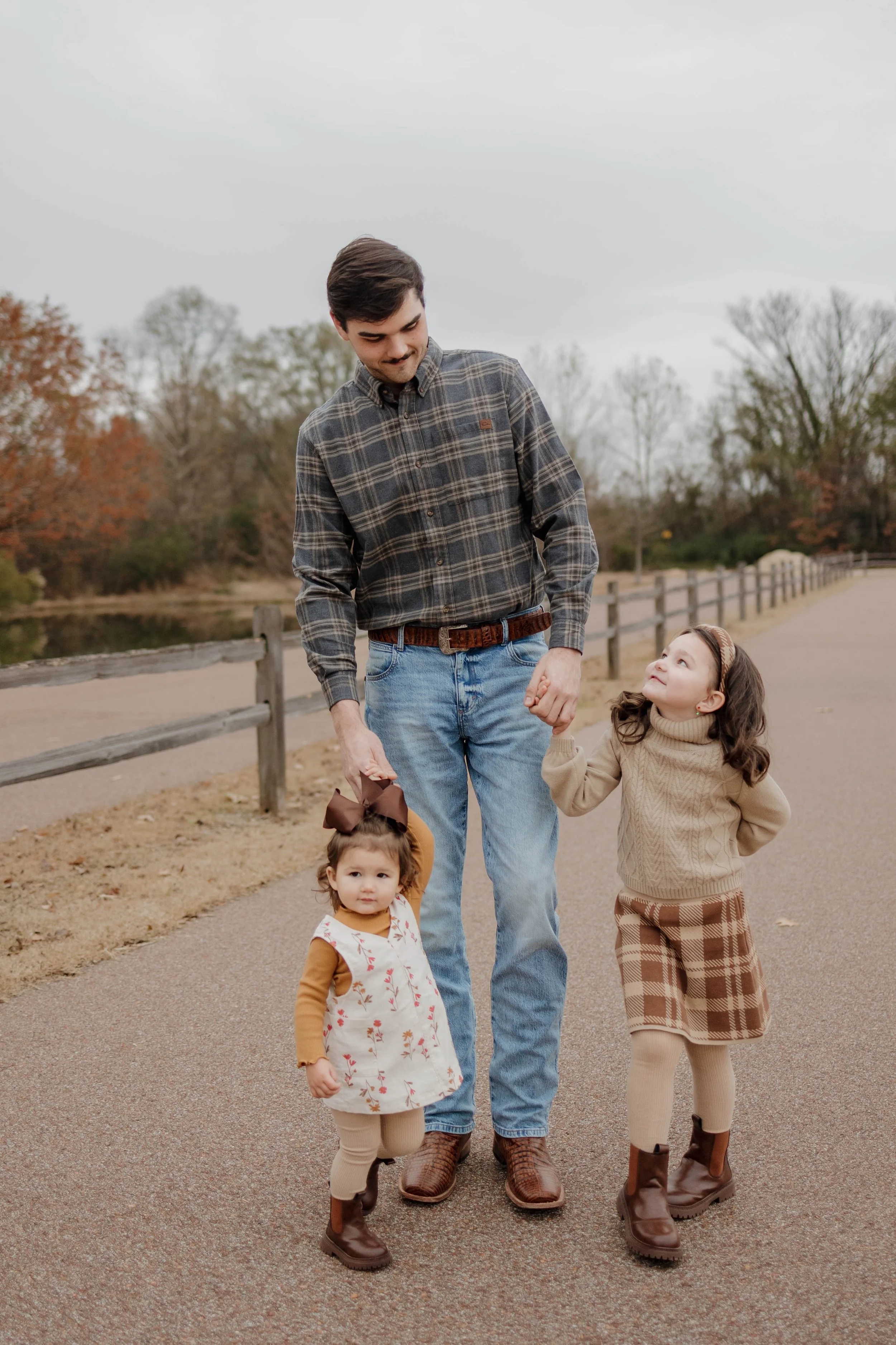 A man walking with three young girls along a paved path in a park during autumn. The man is holding hands with one girl while another girl walks nearby. All are dressed in fall clothing and boots.