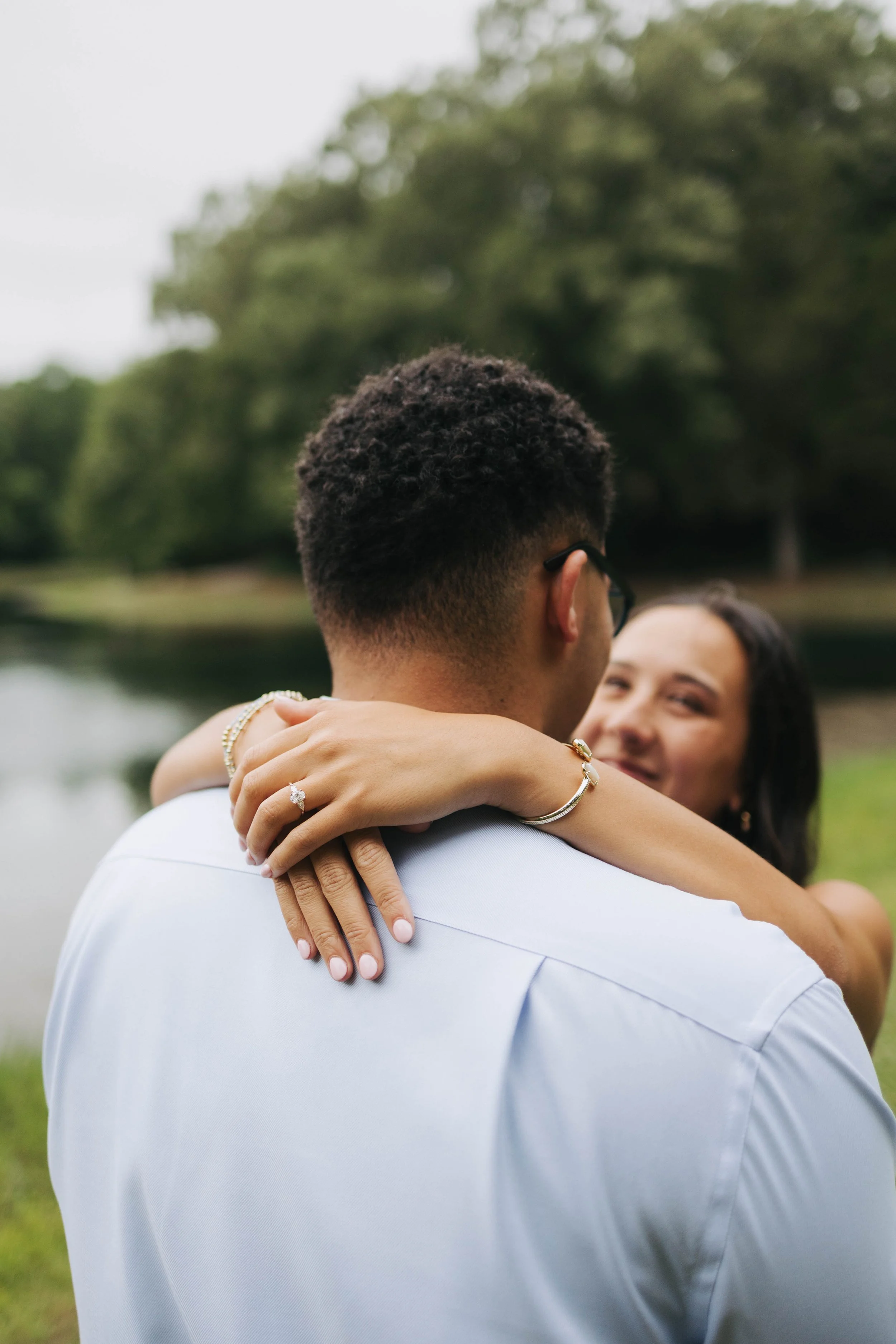 A couple hugging outdoors by a lake with trees in the background. The man has short curly hair and wears glasses, while the woman has dark hair and is smiling.