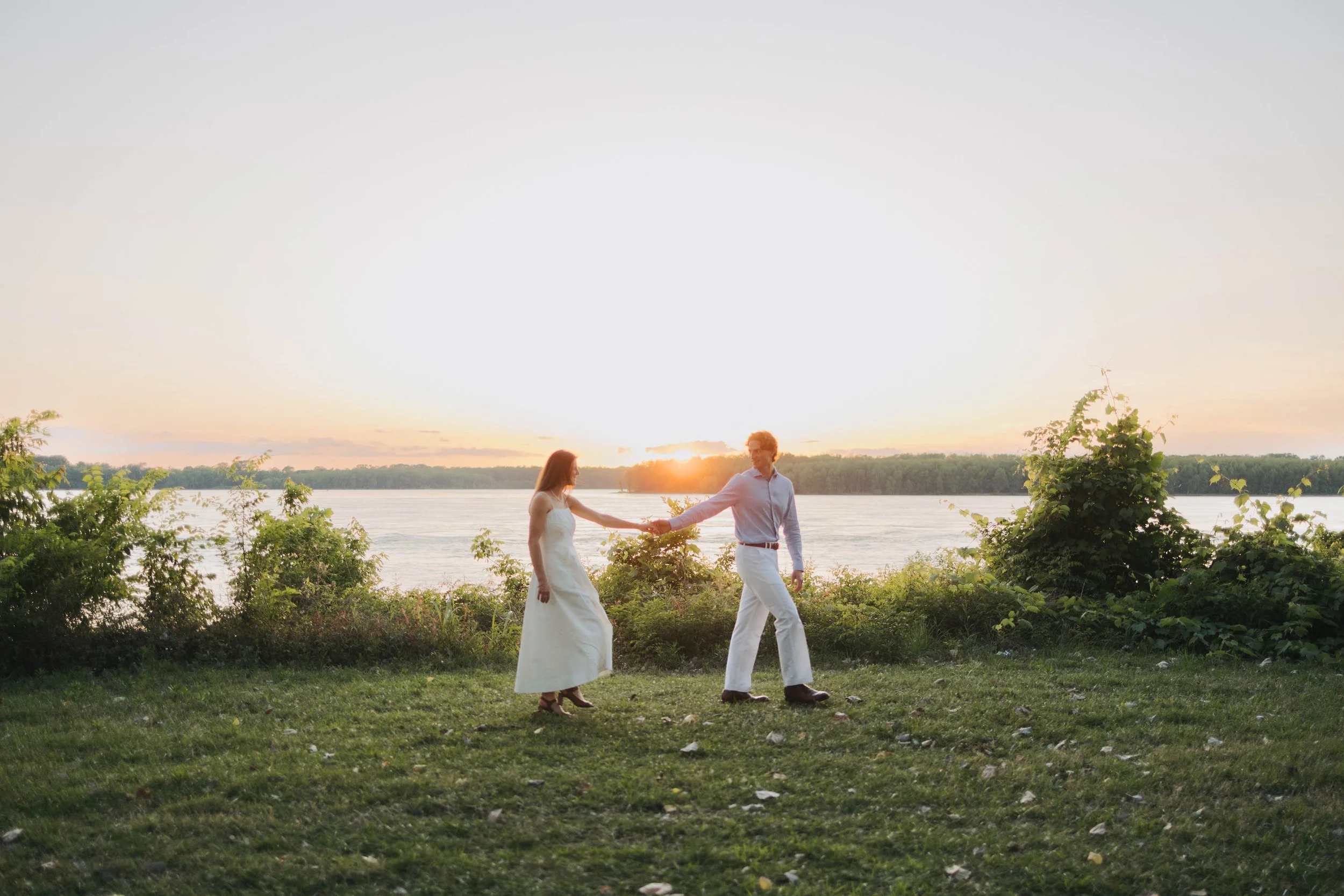 A couple in formal attire holding hands while walking along a grassy area near a body of water during sunset, with trees in the background.