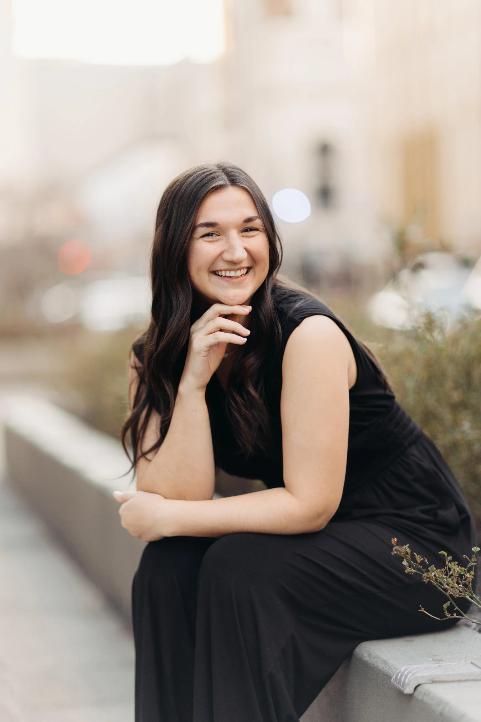 Young woman with long brown hair smiling and sitting outdoors on a concrete ledge, wearing a black sleeveless dress, with a blurred background of city buildings.