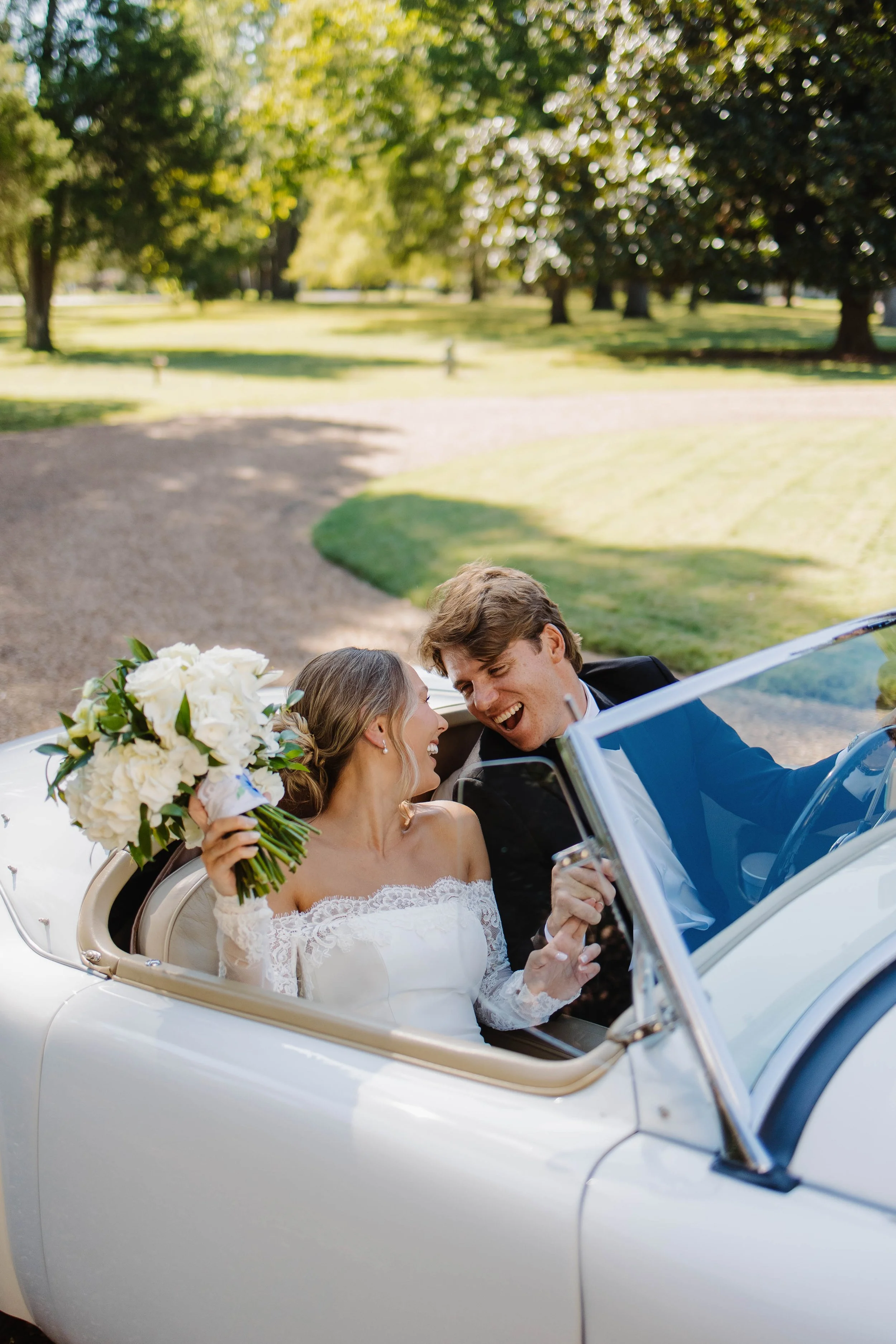 Happy newlywed couple in a vintage convertible car during a wedding celebration in a park with green trees and sunlight.