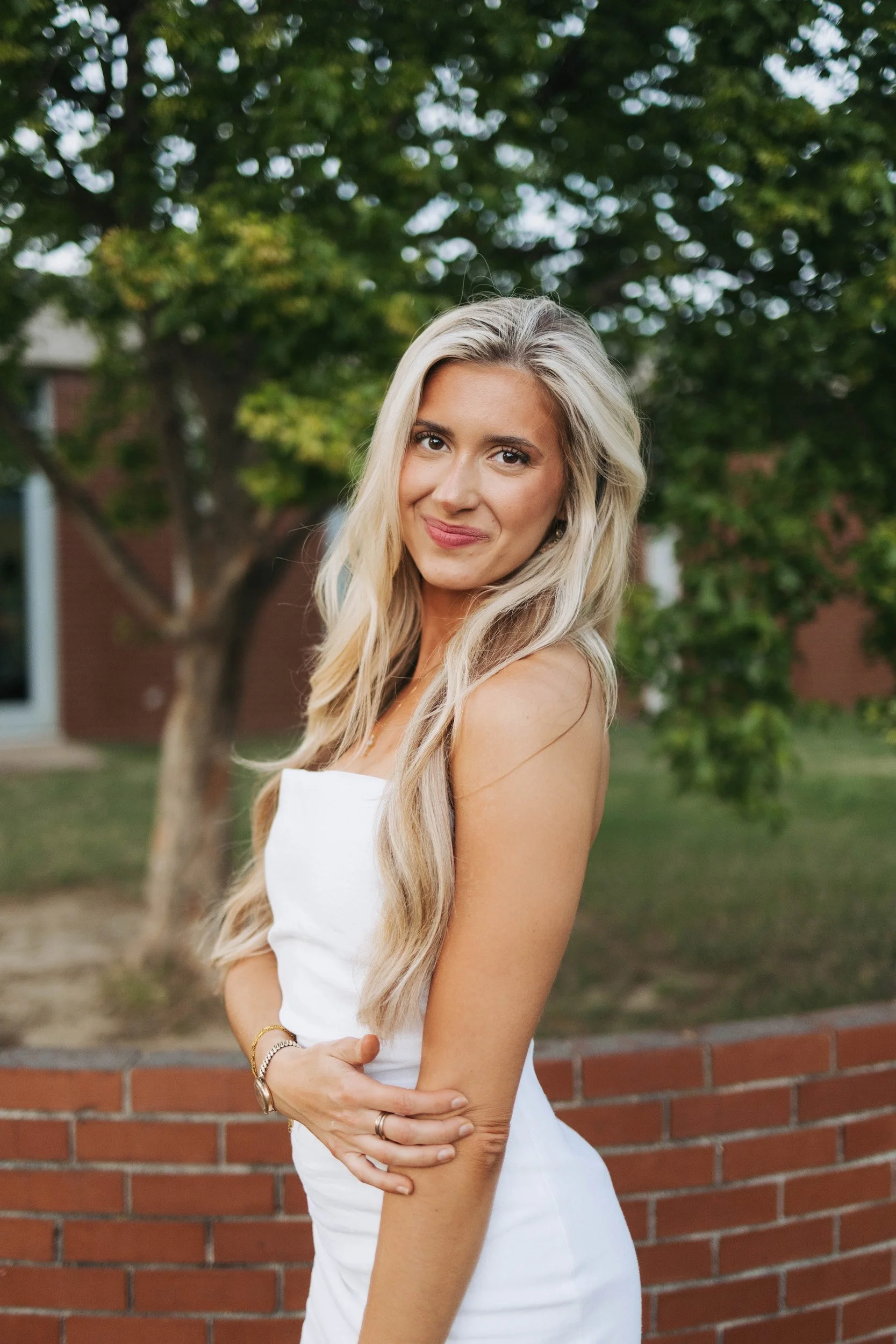 A woman with long blonde wavy hair stands outdoors in front of a brick wall and green trees, smiling softly.