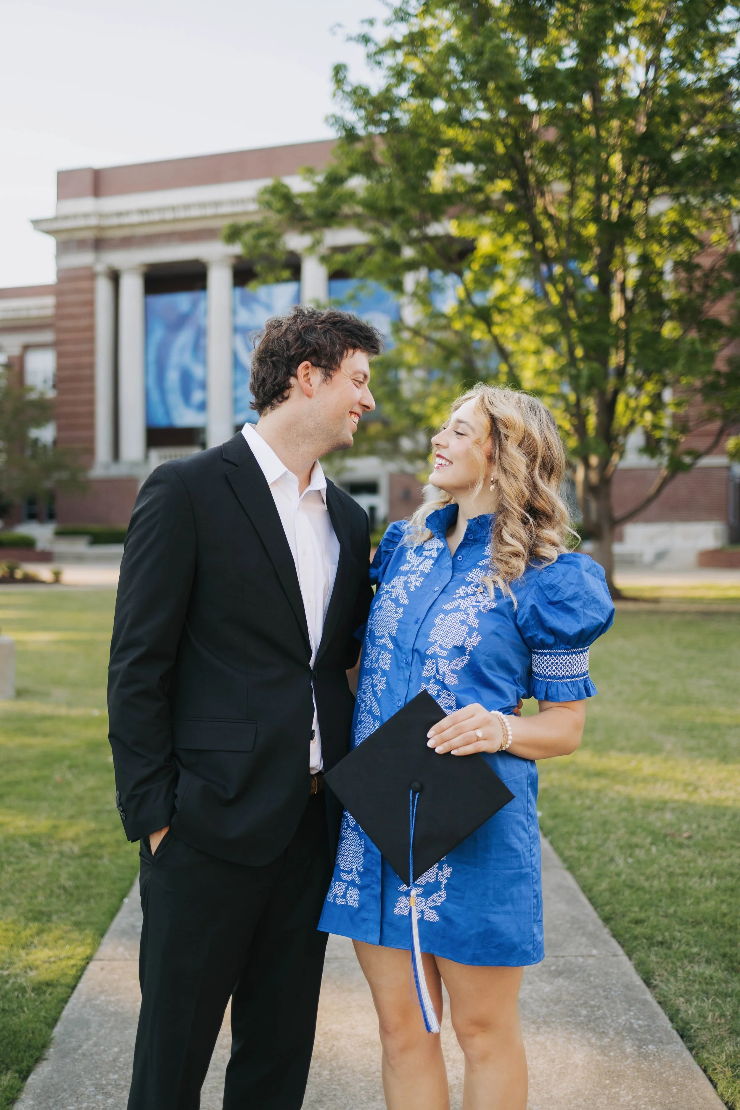 A young man in a suit and a young woman in a blue dress with a graduation cap, standing outdoors on a sidewalk in front of a college building, smiling at each other.