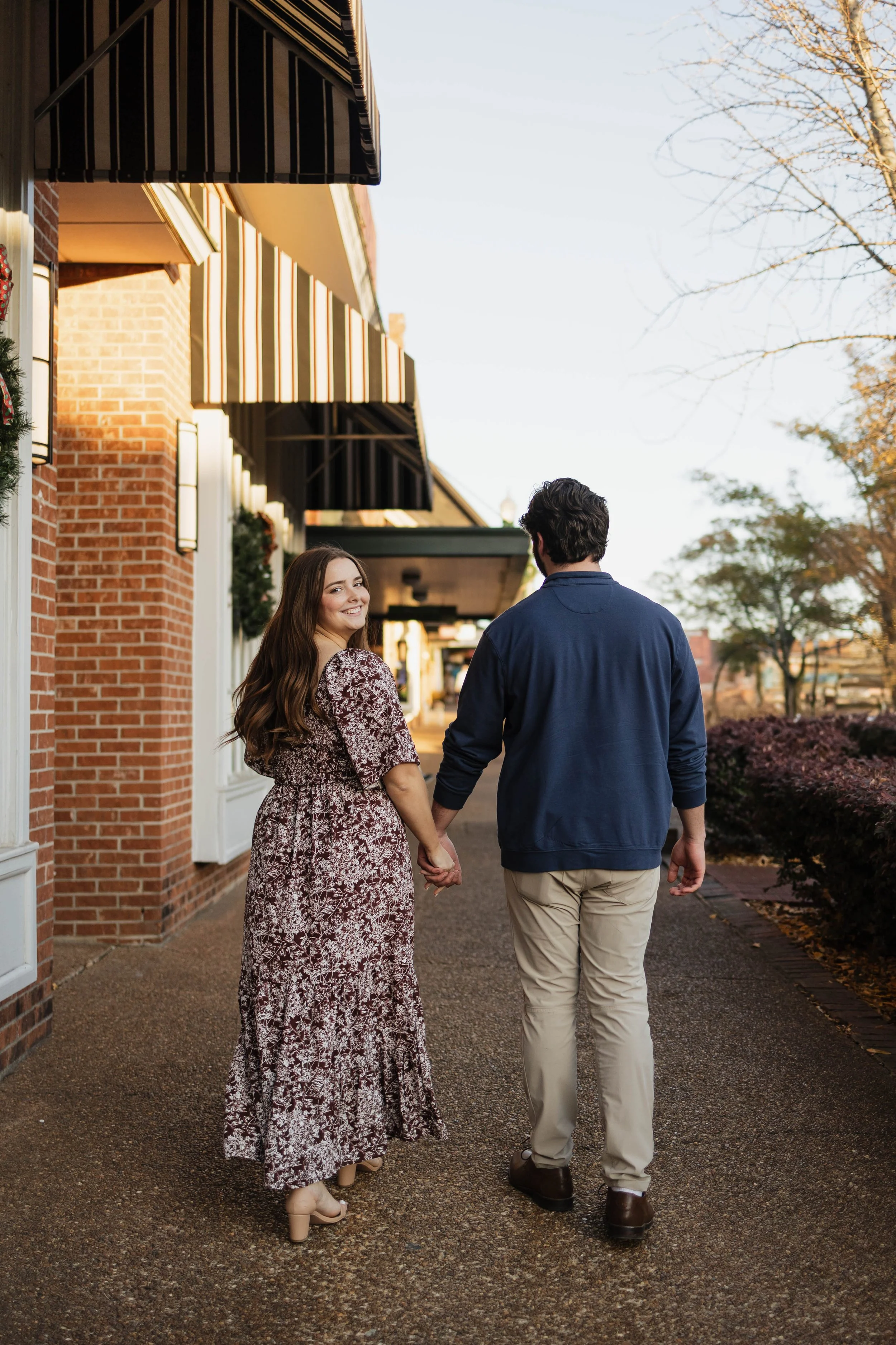A couple holding hands and walking down a sidewalk outside a brick building with striped awnings, decorated for the holidays in the autumn.