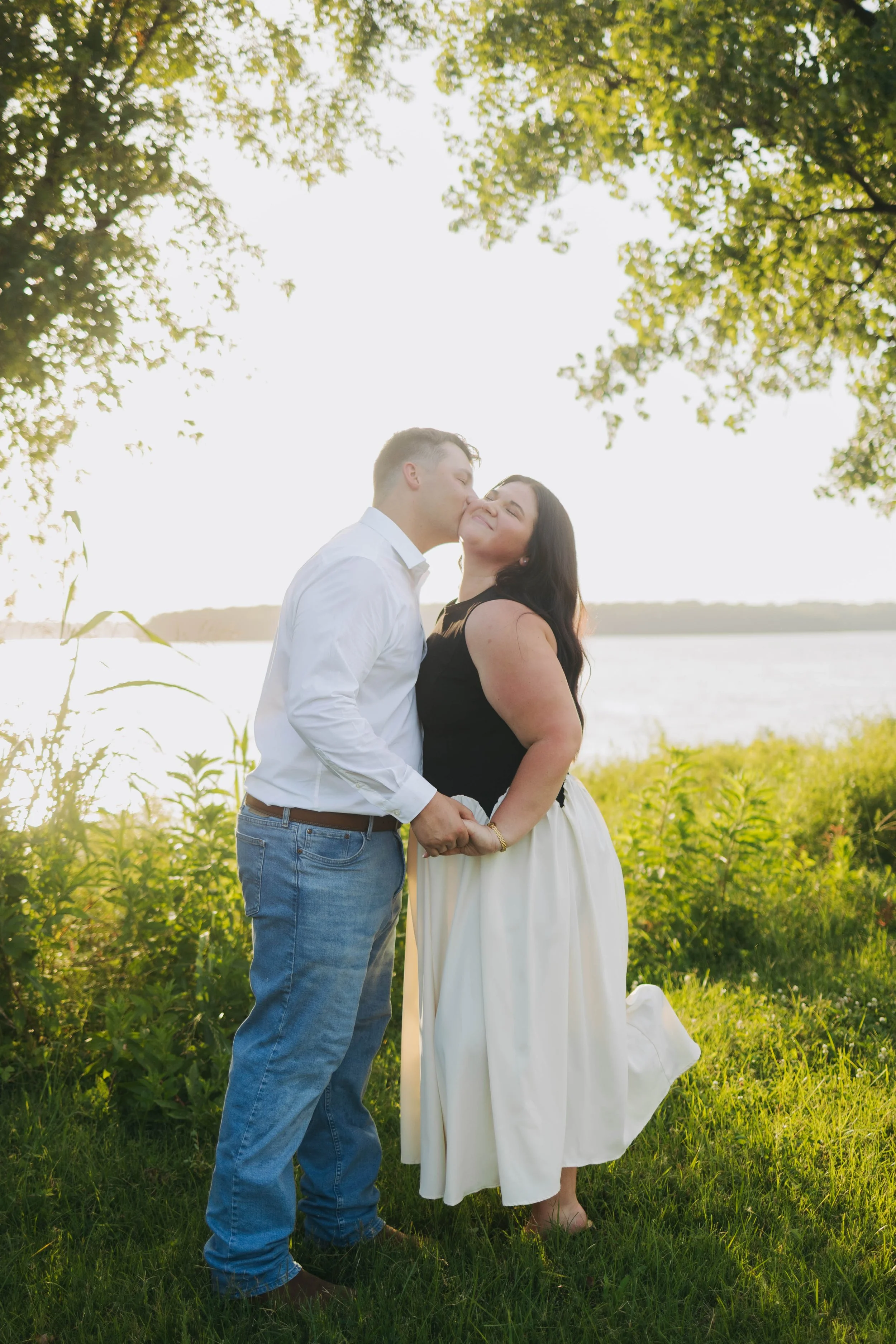 A couple standing outdoors by a river, holding hands, with their eyes closed and smiling, surrounded by green plants and trees, during a sunny day.