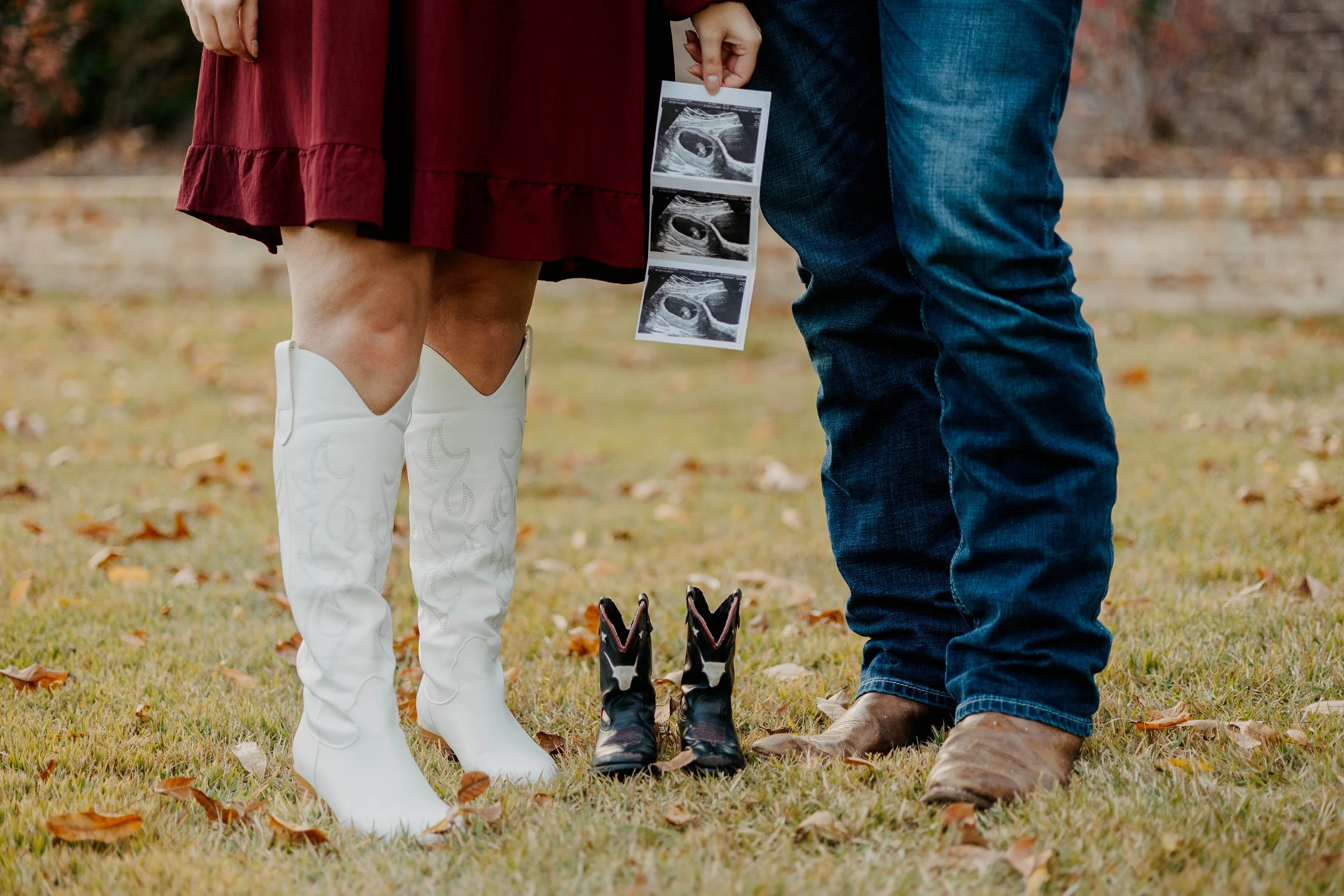 A pregnant woman in white cowboy boots holding ultrasound pictures standing next to a man in jeans and brown boots, as they stand outdoors on grass covered with fallen autumn leaves.