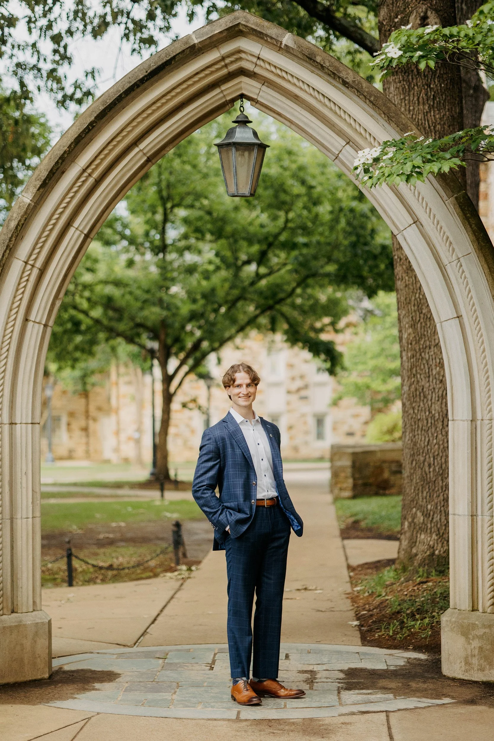 A man in a blue checkered suit with a white shirt and brown leather shoes standing under a stone archway with a hanging lantern, green trees in the background.