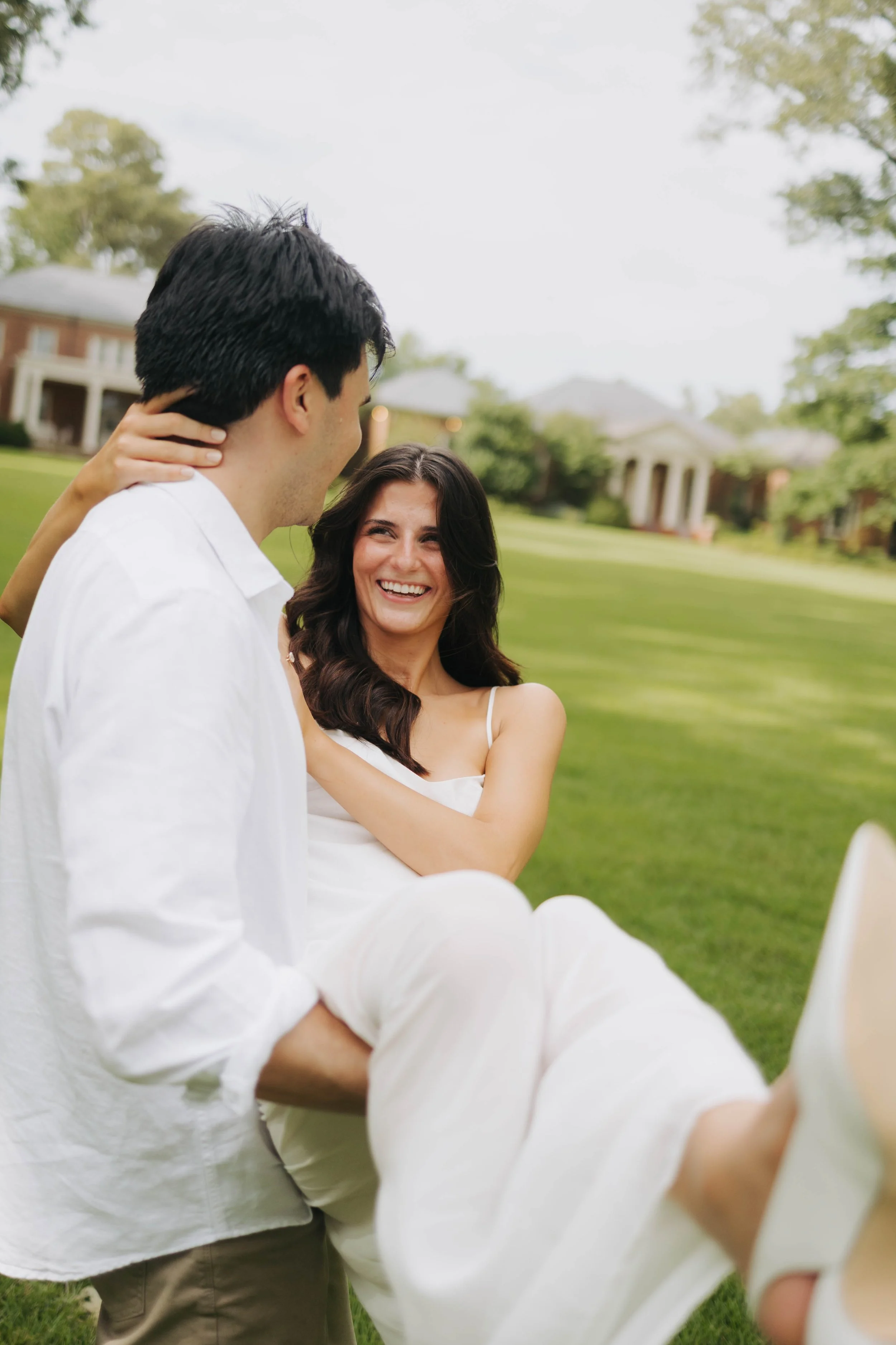 A man with dark hair, wearing a white shirt, and a woman with long dark hair, smiling and wearing a white dress, are outdoors in a grassy area with trees and houses in the background. The man is looking at the woman and holding her around the waist, 