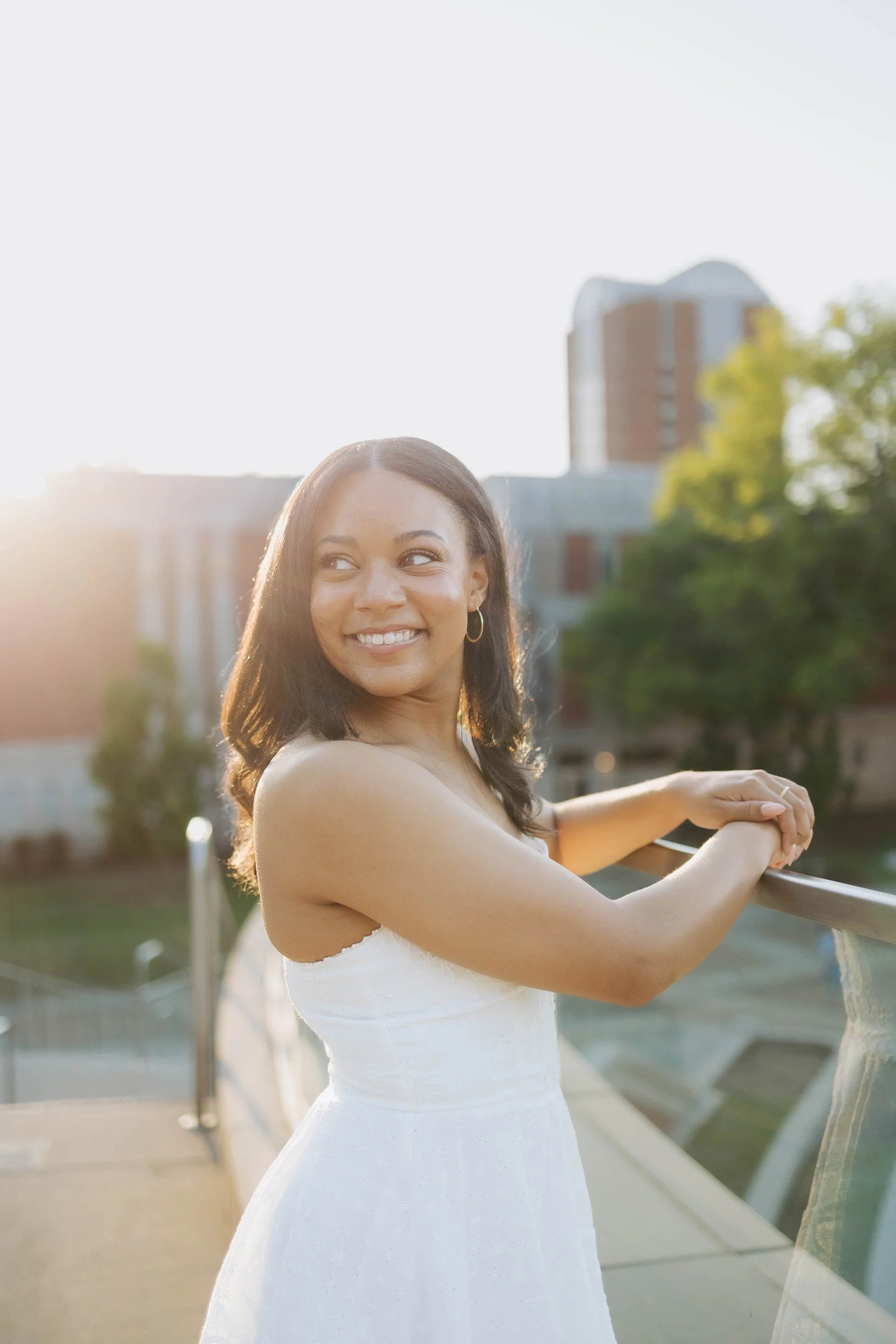 A young woman in a white dress standing outdoors on a bridge, smiling, with sunlight shining behind her and a building in the background.