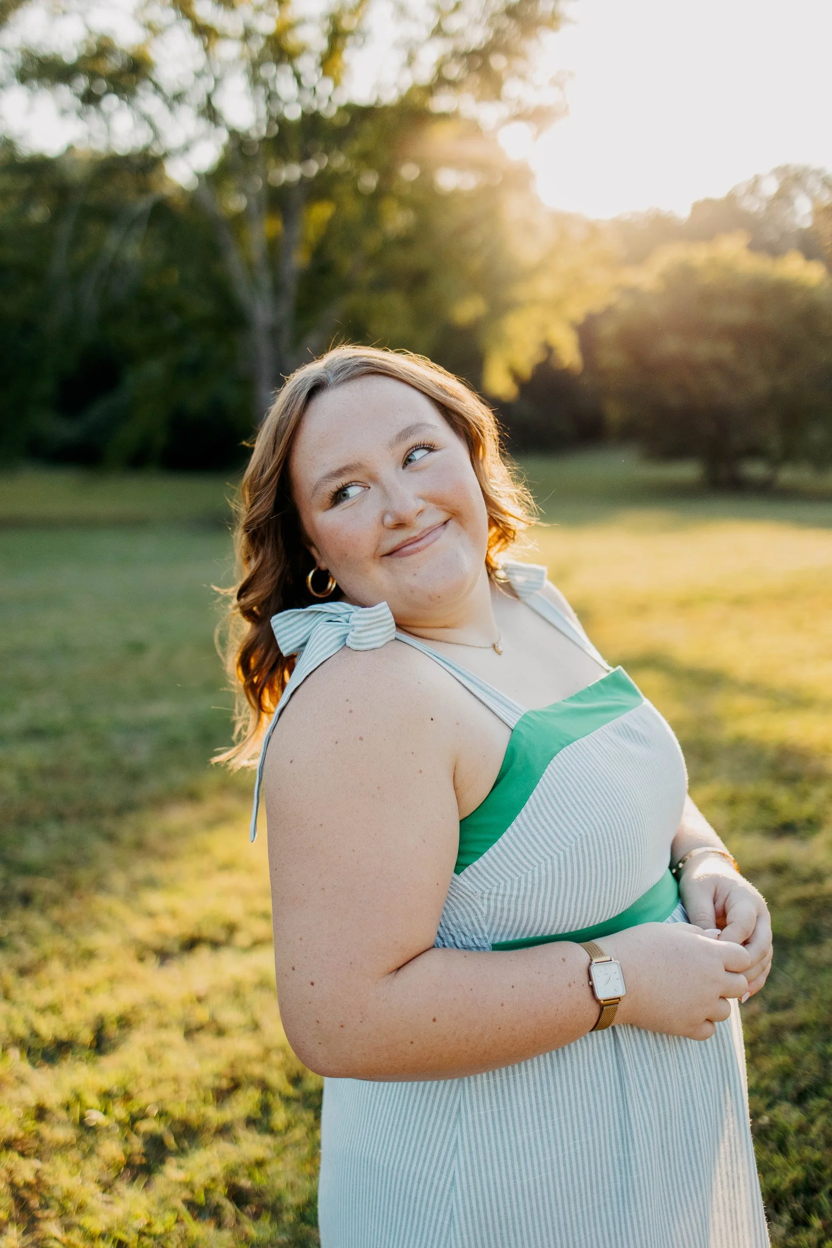 A woman with red hair and freckles smiling outdoors during golden hour, wearing a sleeveless dress with green and white stripes, standing in a grassy area with trees and sunlight in the background.