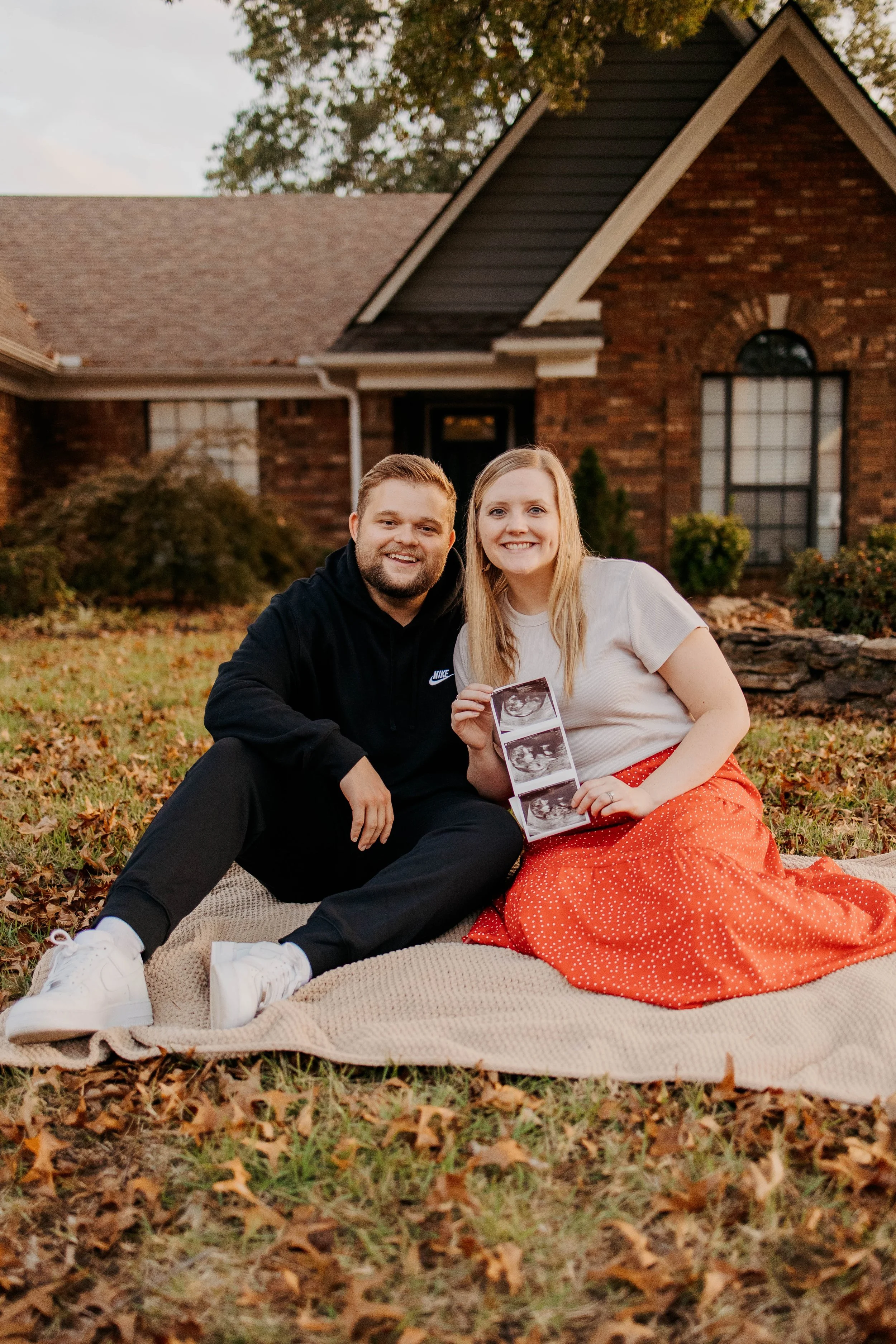 A couple sitting on a blanket outdoors in front of a brick house. The woman is holding ultrasound photo strips. They are smiling, and the woman is wearing a white top and a red skirt. The man is wearing a black hoodie and black pants. There are autum