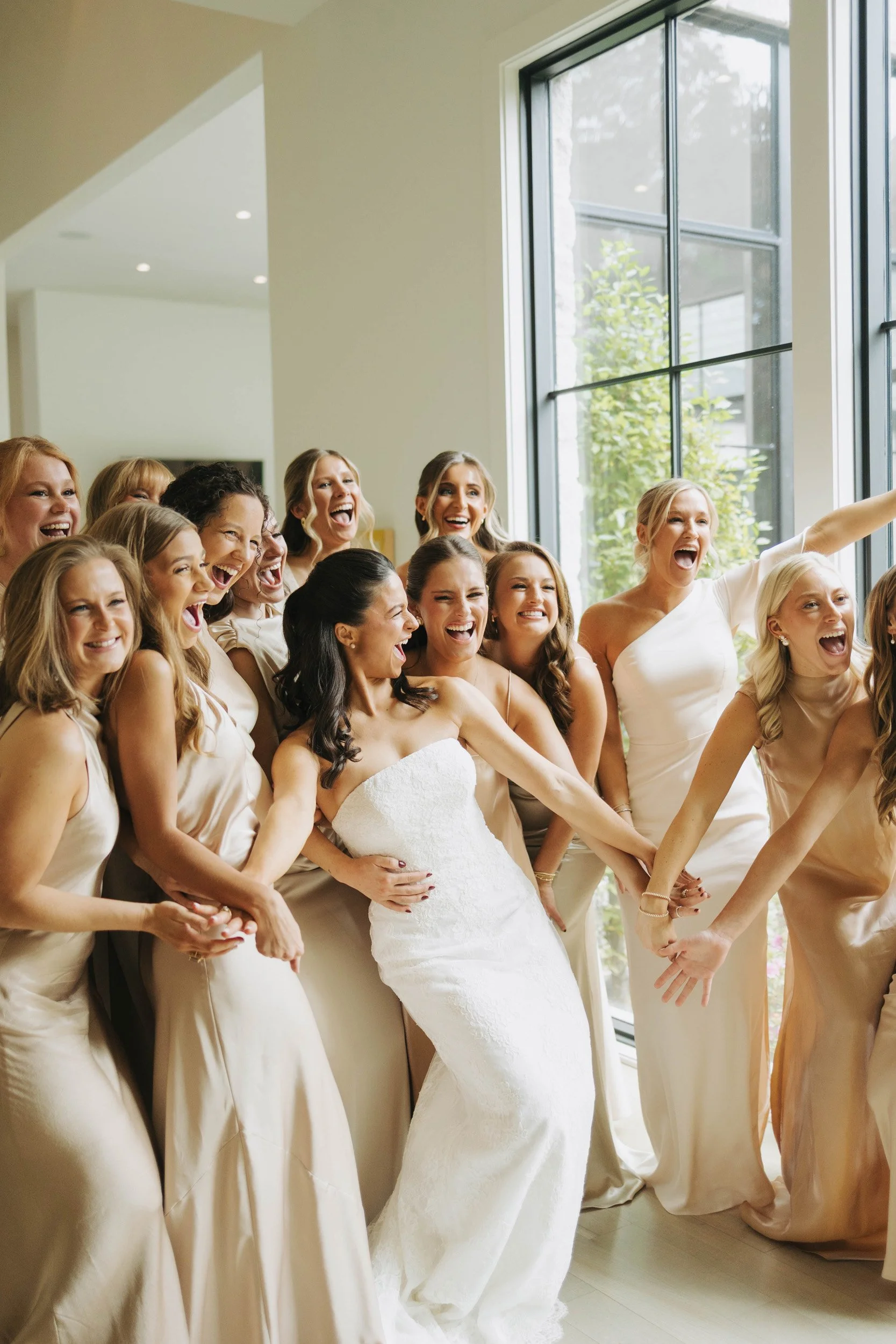 A group of women, including a bride in a white wedding dress, celebrating and smiling together by a large window.