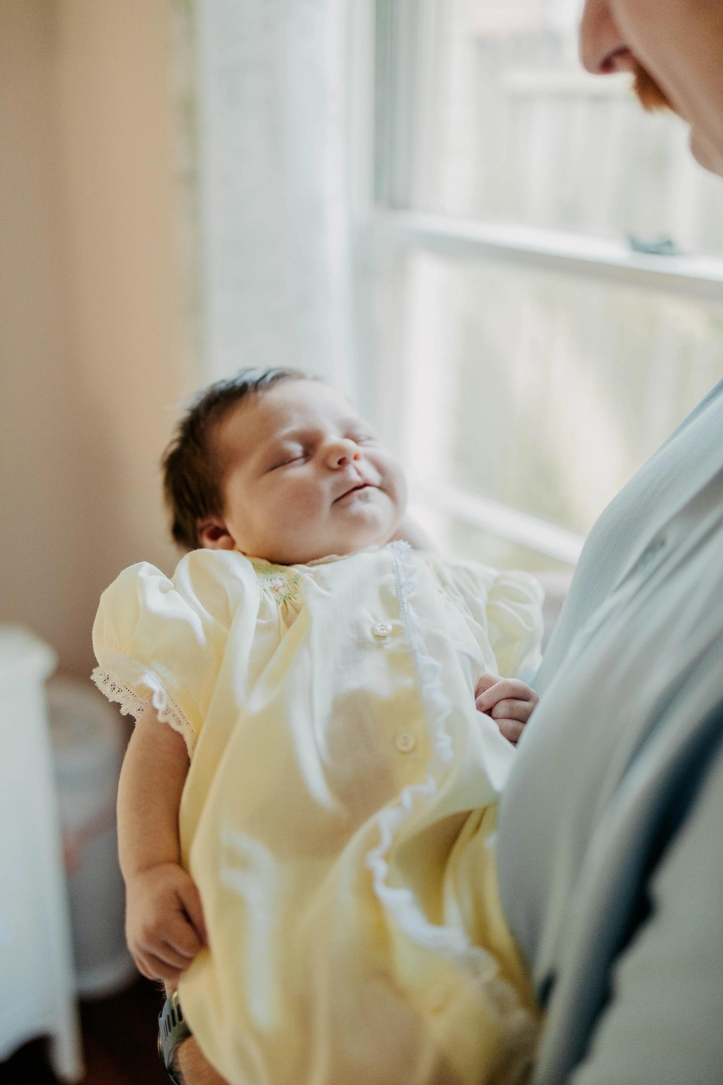 A young baby girl with dark hair, wearing a yellow dress with lace trim, is being held by a man, possibly her father, who is visible from his shoulder to his nose, wearing a light-colored shirt. They are near a window with natural light.