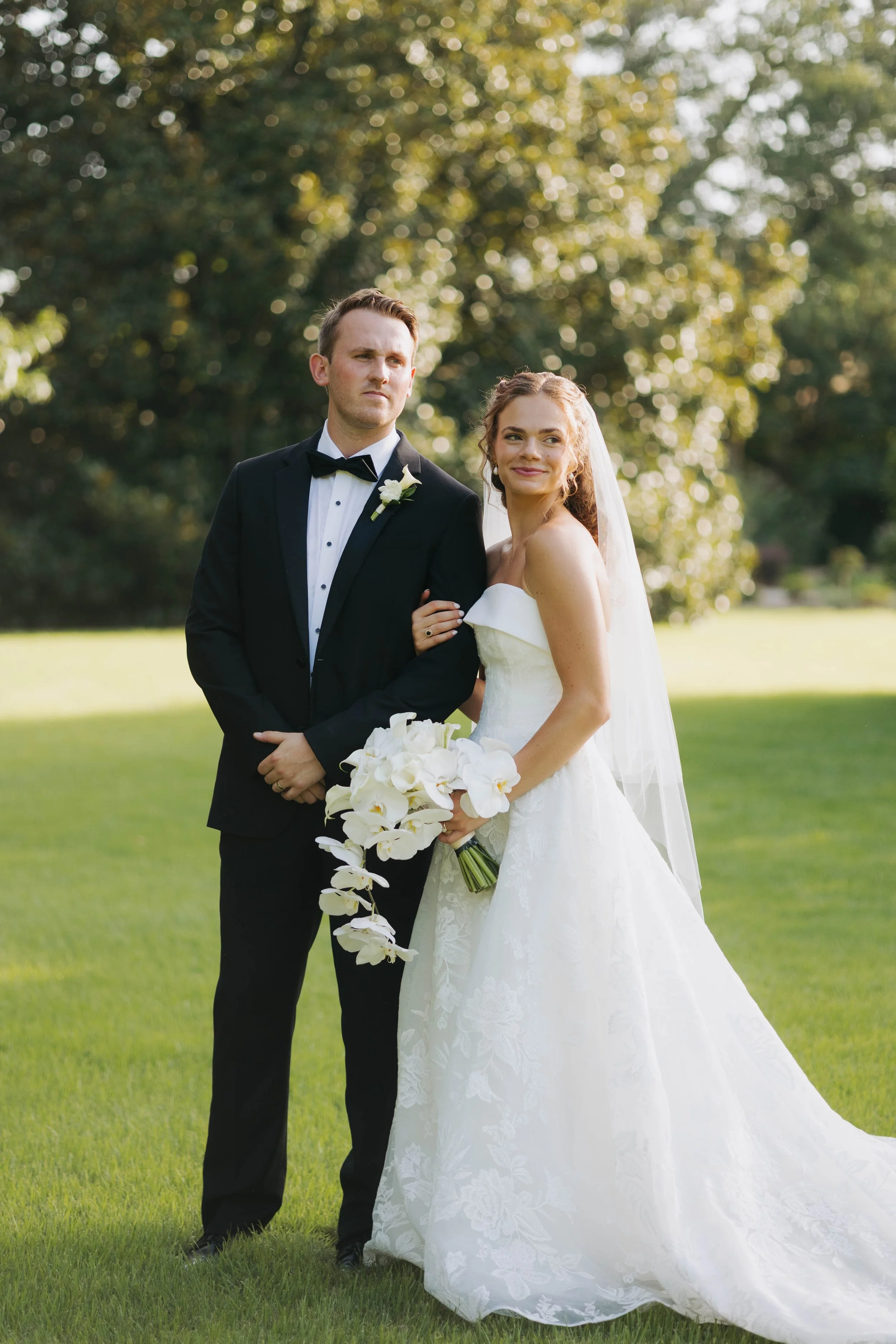 A bride and groom stand outdoors on a grassy field, the groom in a black tuxedo and the bride in a white strapless wedding gown holding a bouquet of white flowers, with trees and sunlight in the background.