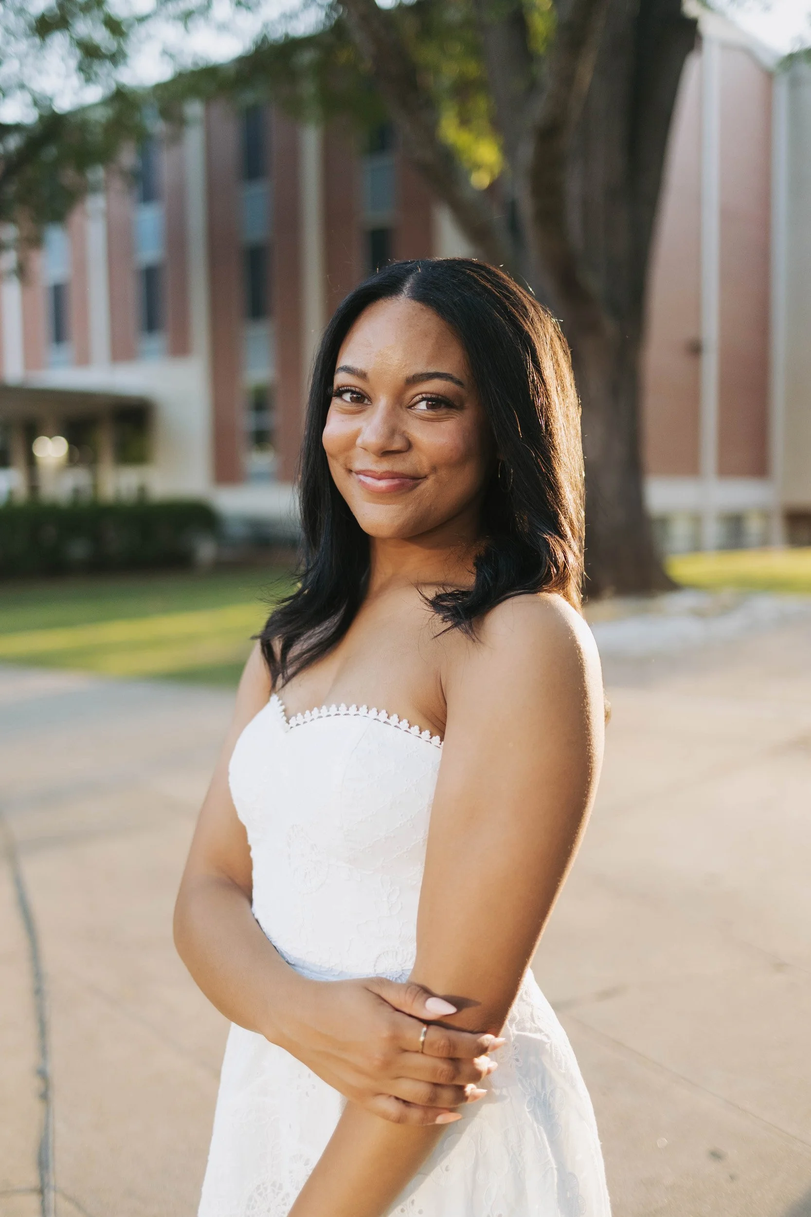 A young woman standing outdoors in a white dress, smiling softly with her arms crossed in front of her. She has long dark hair and is near a large tree with a brick building in the background during the daytime.