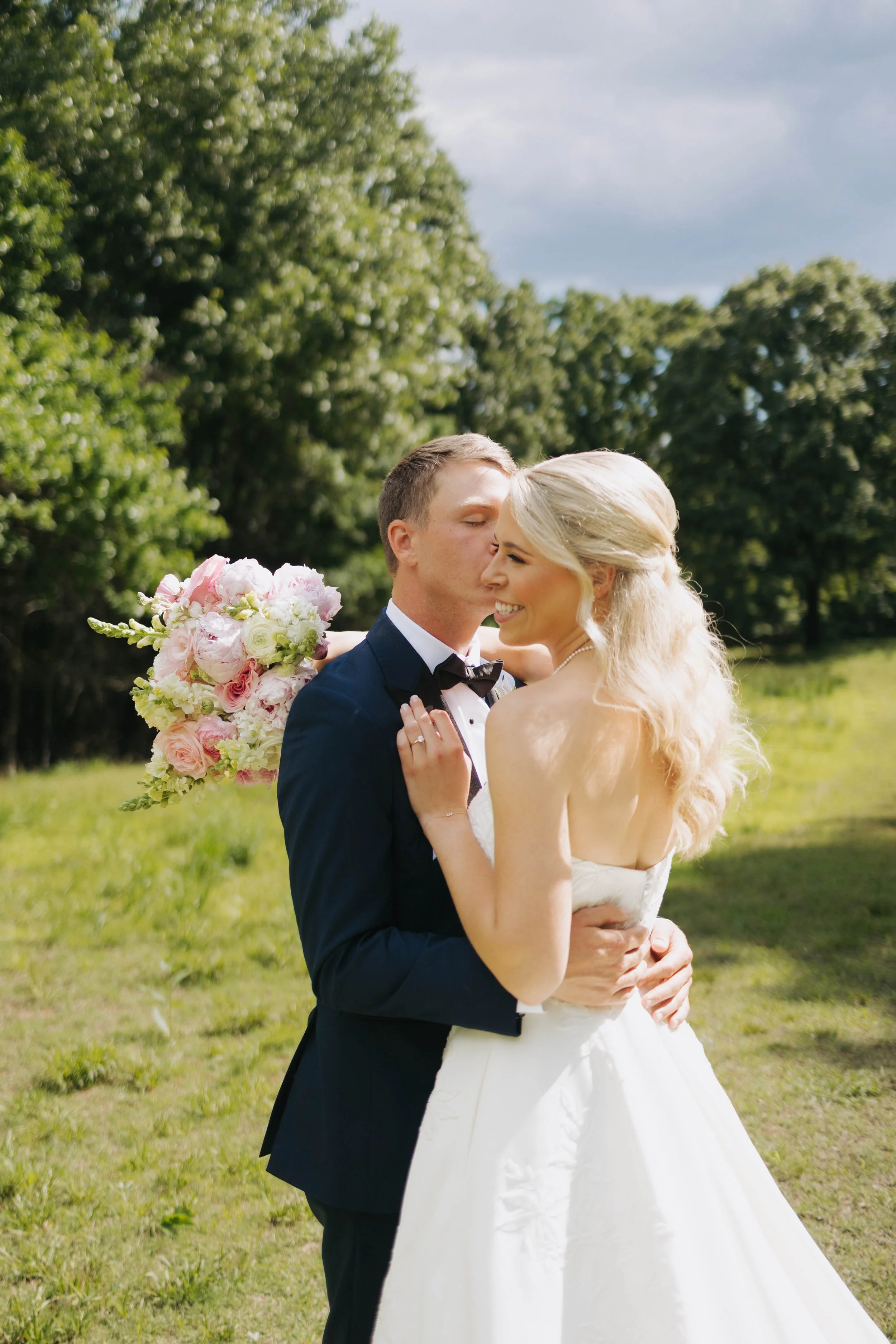 A newlywed couple dressed in wedding attire, hugging outdoors on a grassy area with trees in the background. The groom, in a black tuxedo with a bow tie, is kissing the bride on her cheek. The bride, in a white wedding dress, is smiling with her eyes