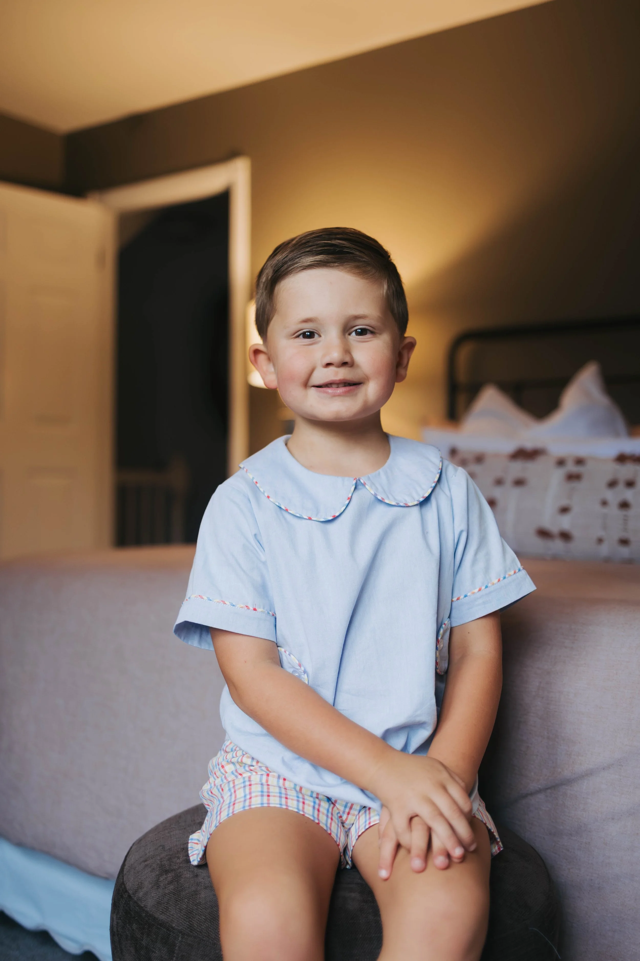 A young boy sitting on a round, dark-colored ottoman in a bedroom, smiling at the camera, wearing a light blue shirt and plaid shorts, with a bed and open closet in the background.