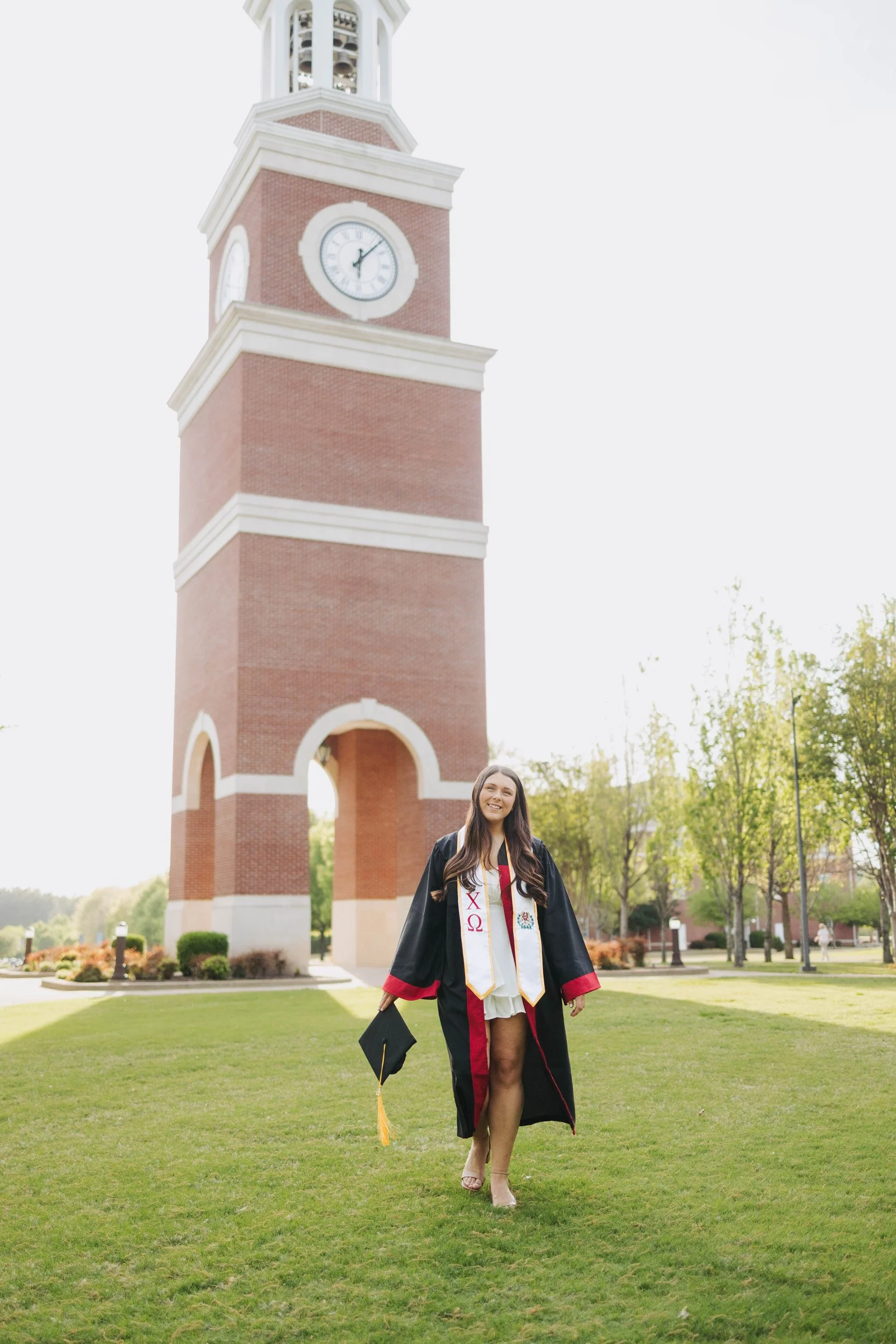 A young woman in a graduation gown and stole standing on a grassy area with a tall brick clock tower in the background. She is holding a mortarboard in her right hand and smiling.