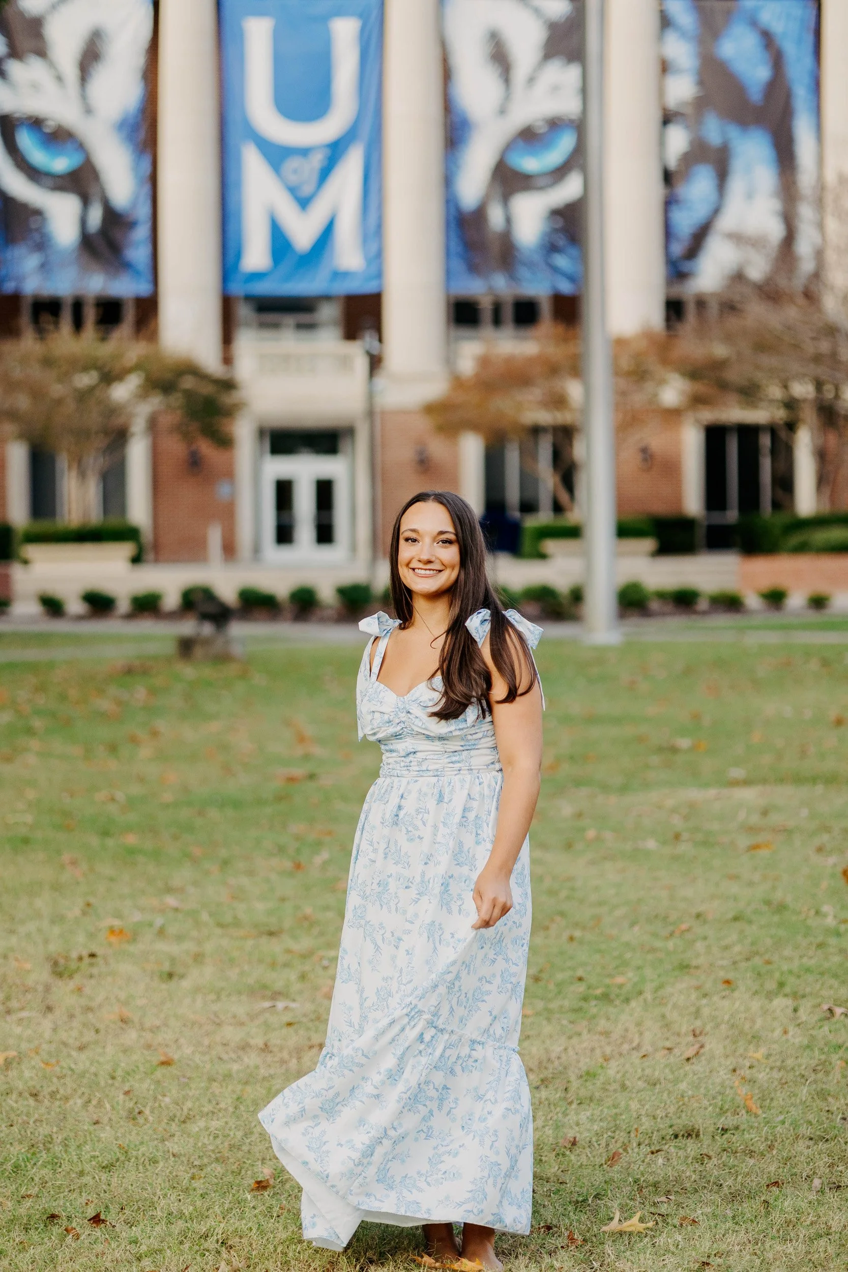 A young woman with long brown hair and a bright smile, wearing a white and blue floral dress, standing outdoors on a grassy area during autumn. In the background, there are flags with a mountain lion face, the letters 'U of M,' and a school building with columns.