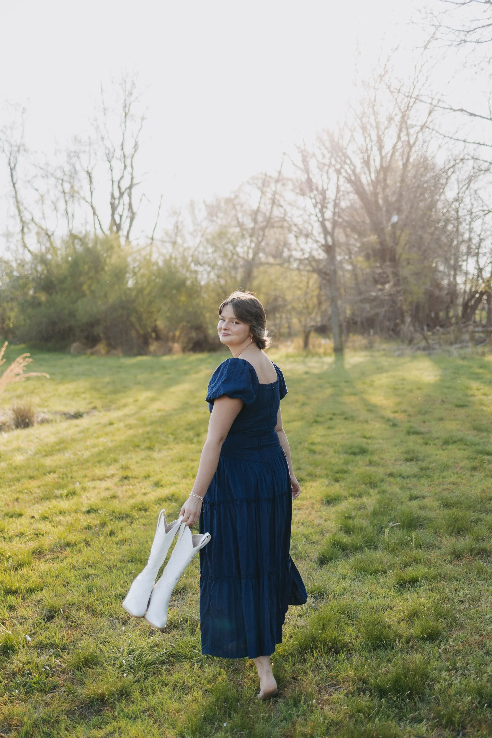 A woman in a long blue dress holds a white bag and walks barefoot across a grassy field in a park with leafless trees in the background and bright sunlight