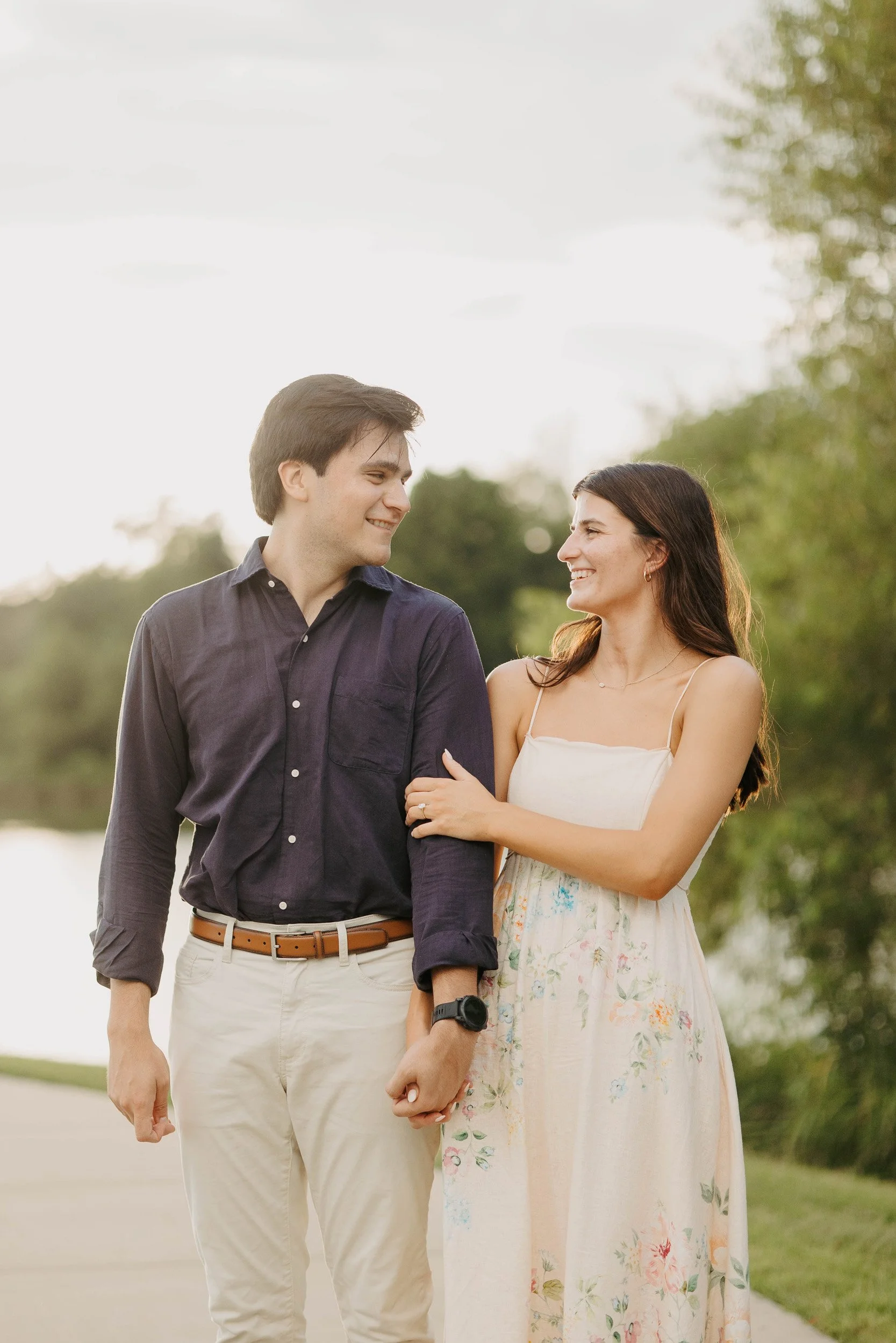 A young man and woman smiling at each other, holding hands outdoors during daytime with trees and greenery in the background.