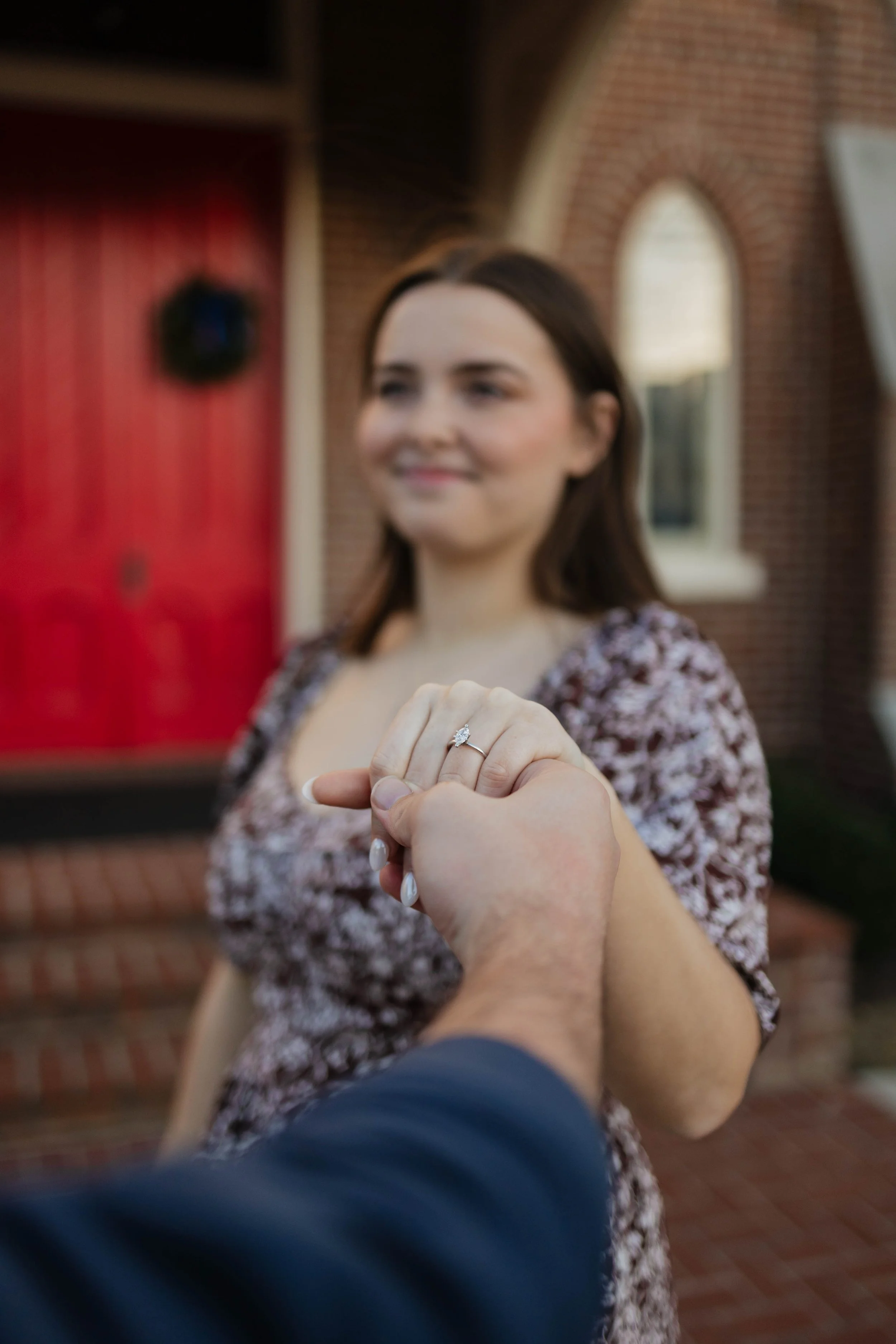 A woman with brown hair smiling while holding hands with someone, displaying an engagement ring. She is standing outside in front of a brick house with large windows and a red door.