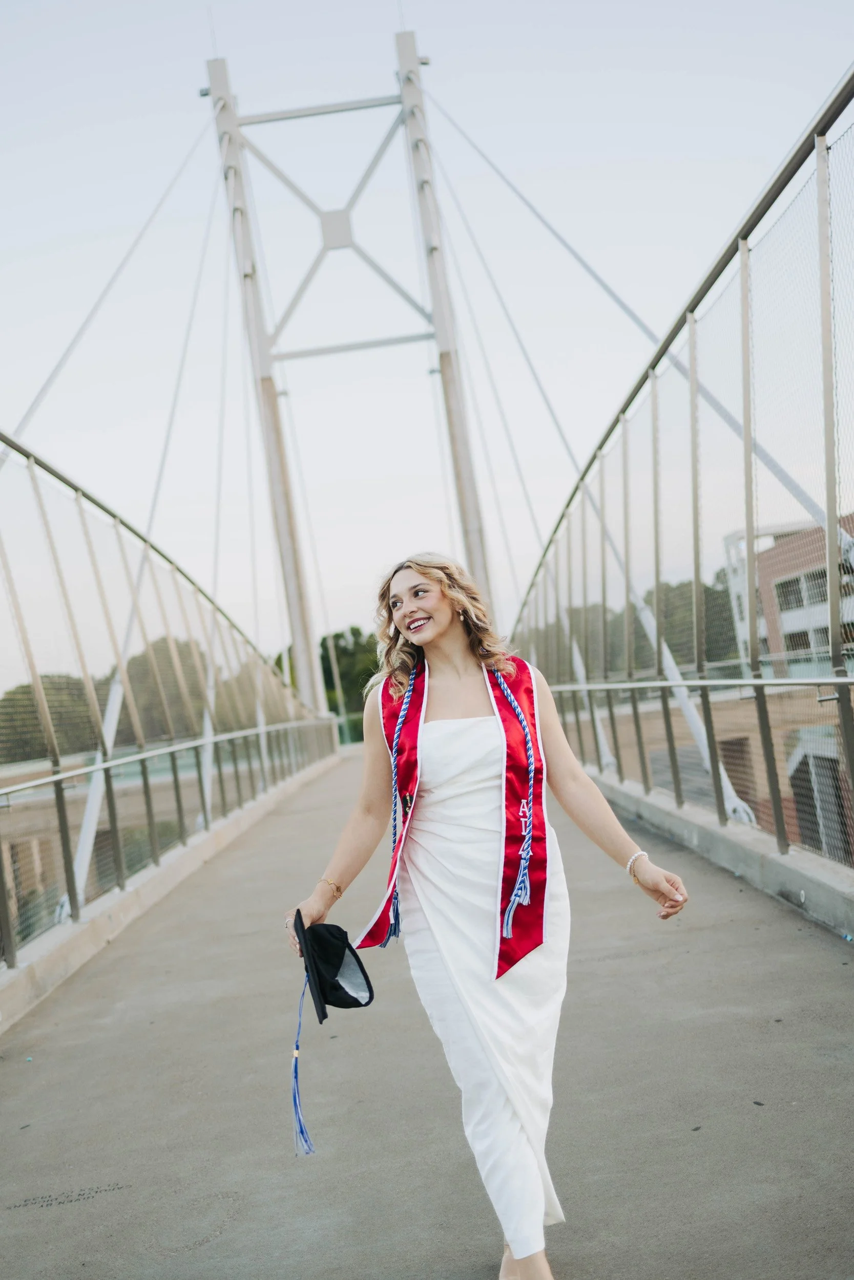 A young woman in a white dress and red sash walks on a bridge, holding a face mask and a black item, smiling.
