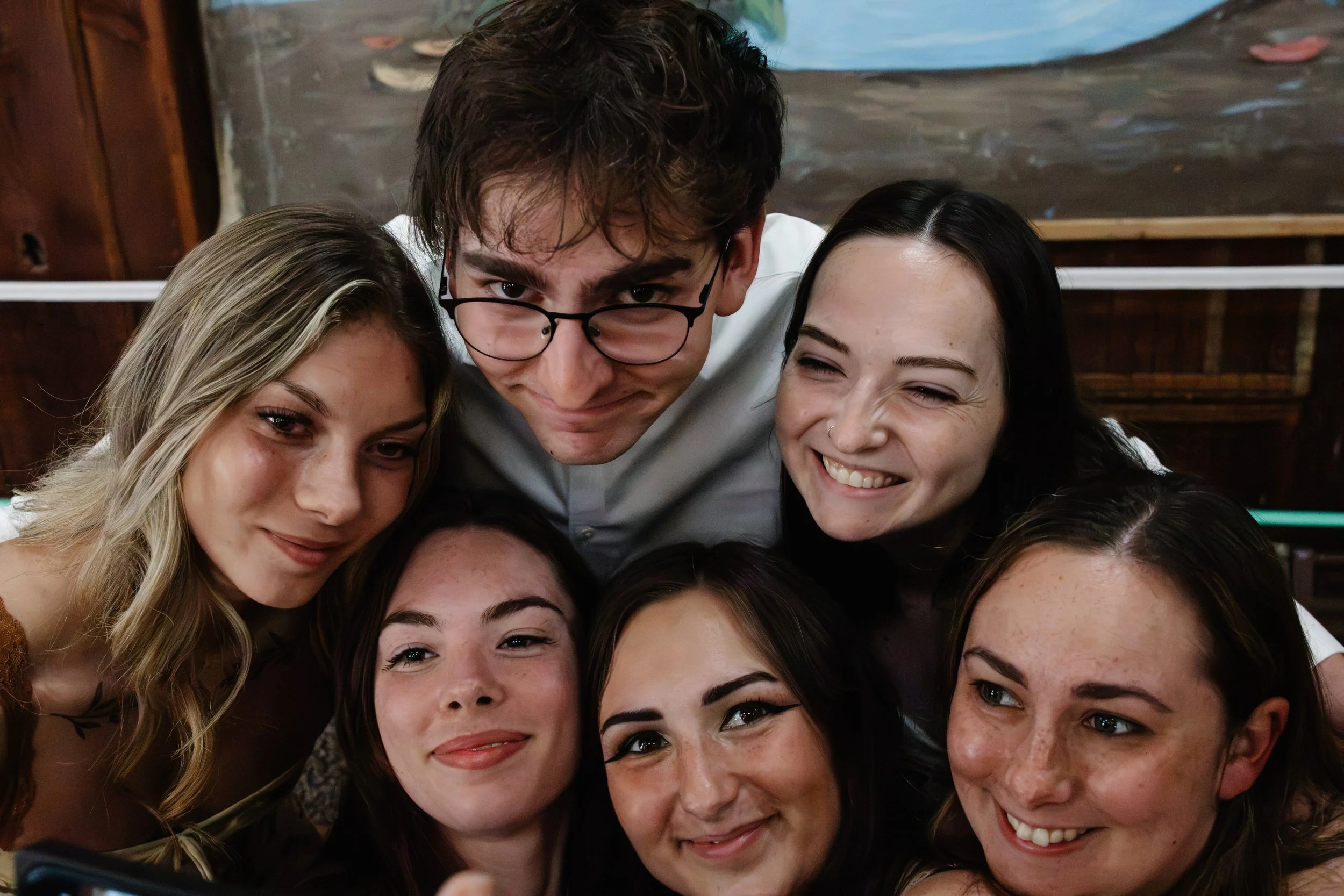 Group of young people taking a close-up selfie together indoors, smiling and showing faces with varying hair colors and styles.