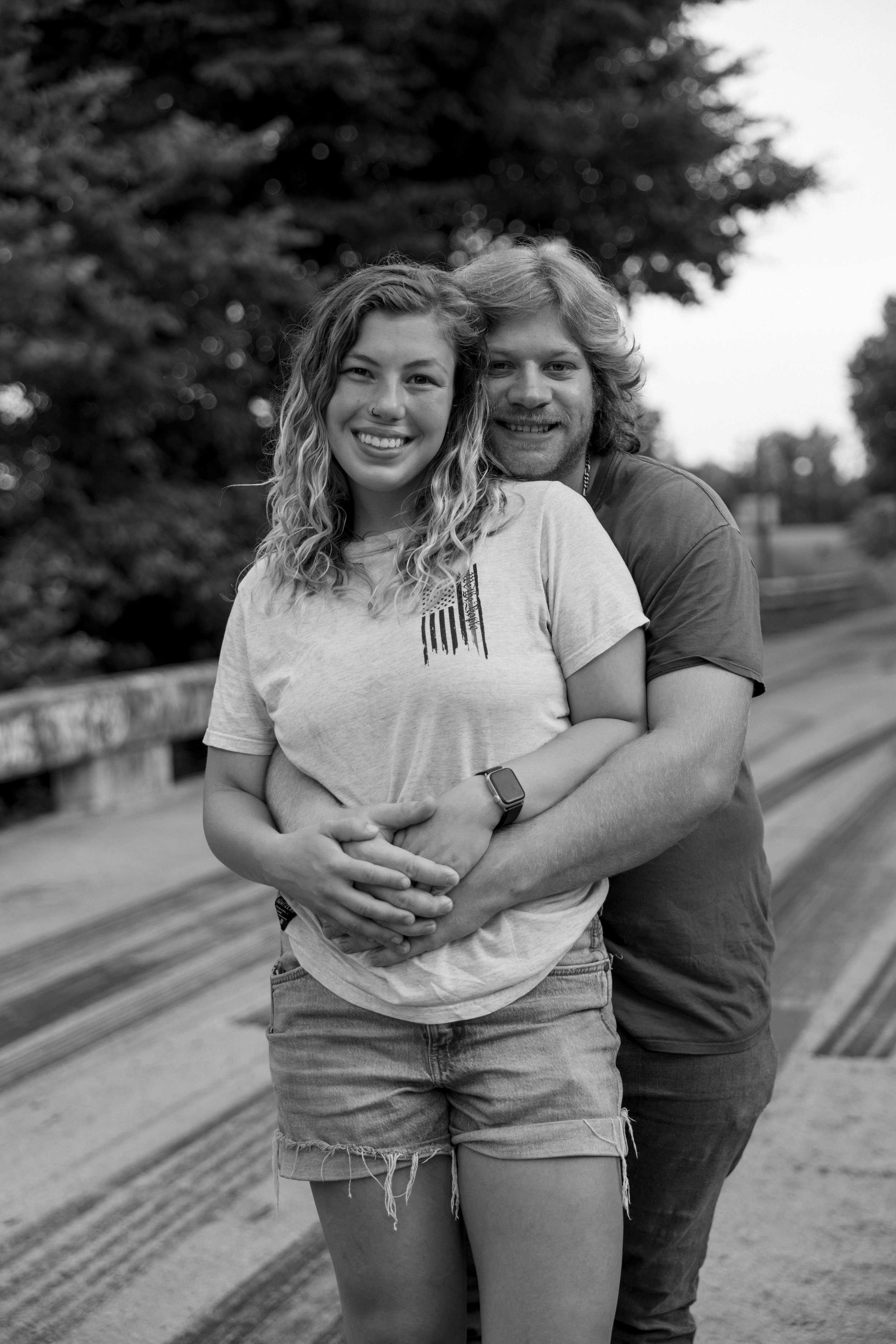 Black and white photo of a young woman with curly hair and a young man with wavy hair smiling and hugging each other outdoors, with trees and a wooden bridge in the background.