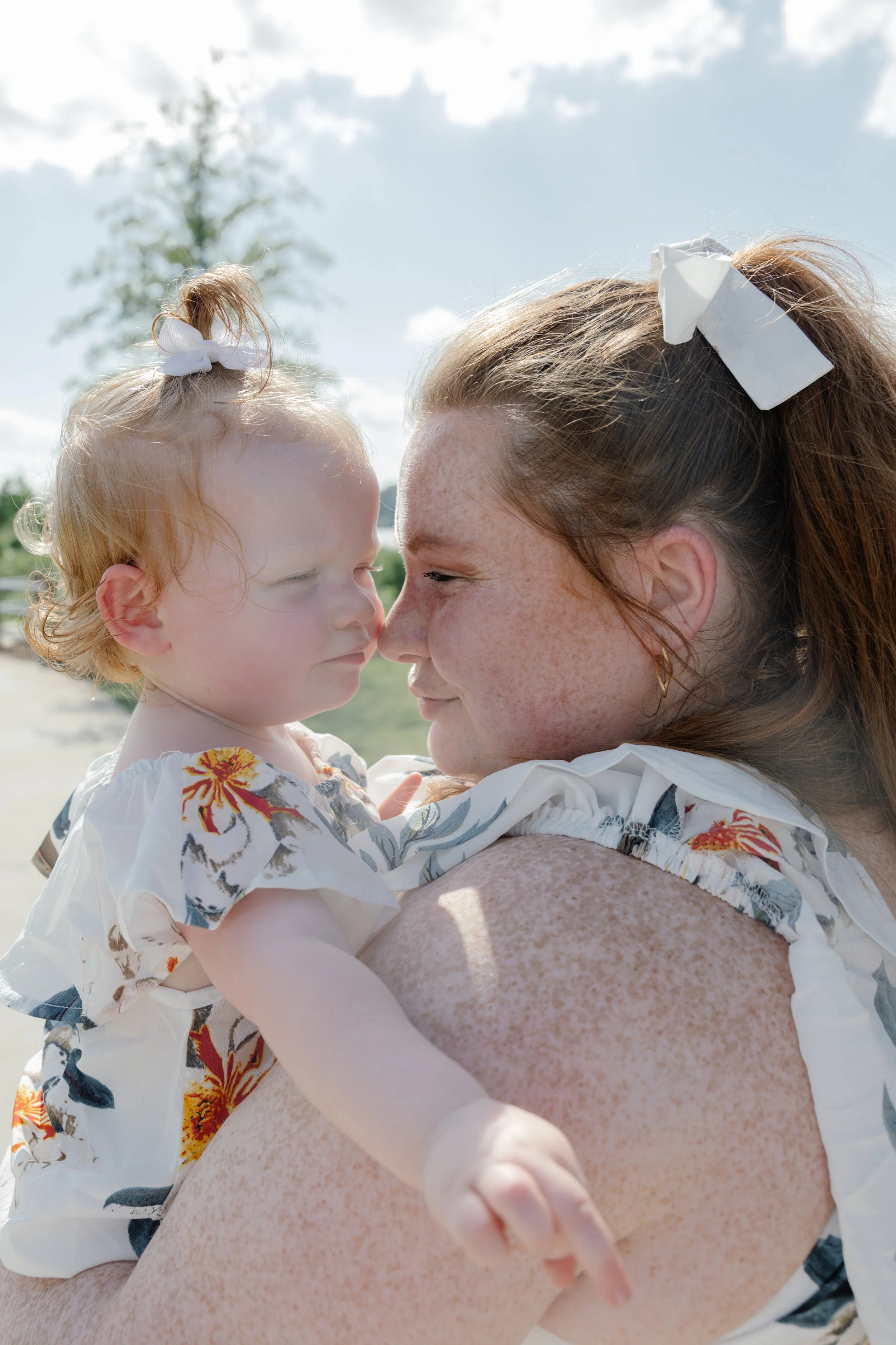 A woman and a young girl are touching foreheads. The woman has red hair with a white bow, and the girl has blonde hair tied with a white bow. They are outdoors on a sunny day.