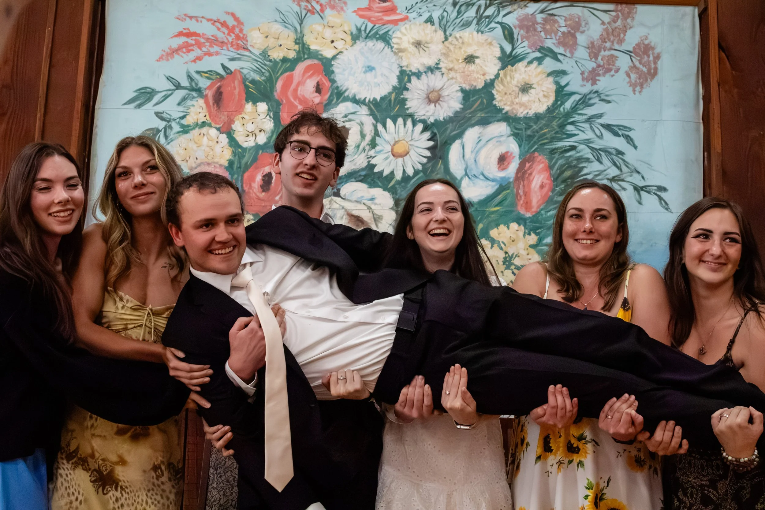 Group of young adults celebrating, with a man in a tuxedo and white shirt in the center, in front of a floral painting backdrop.