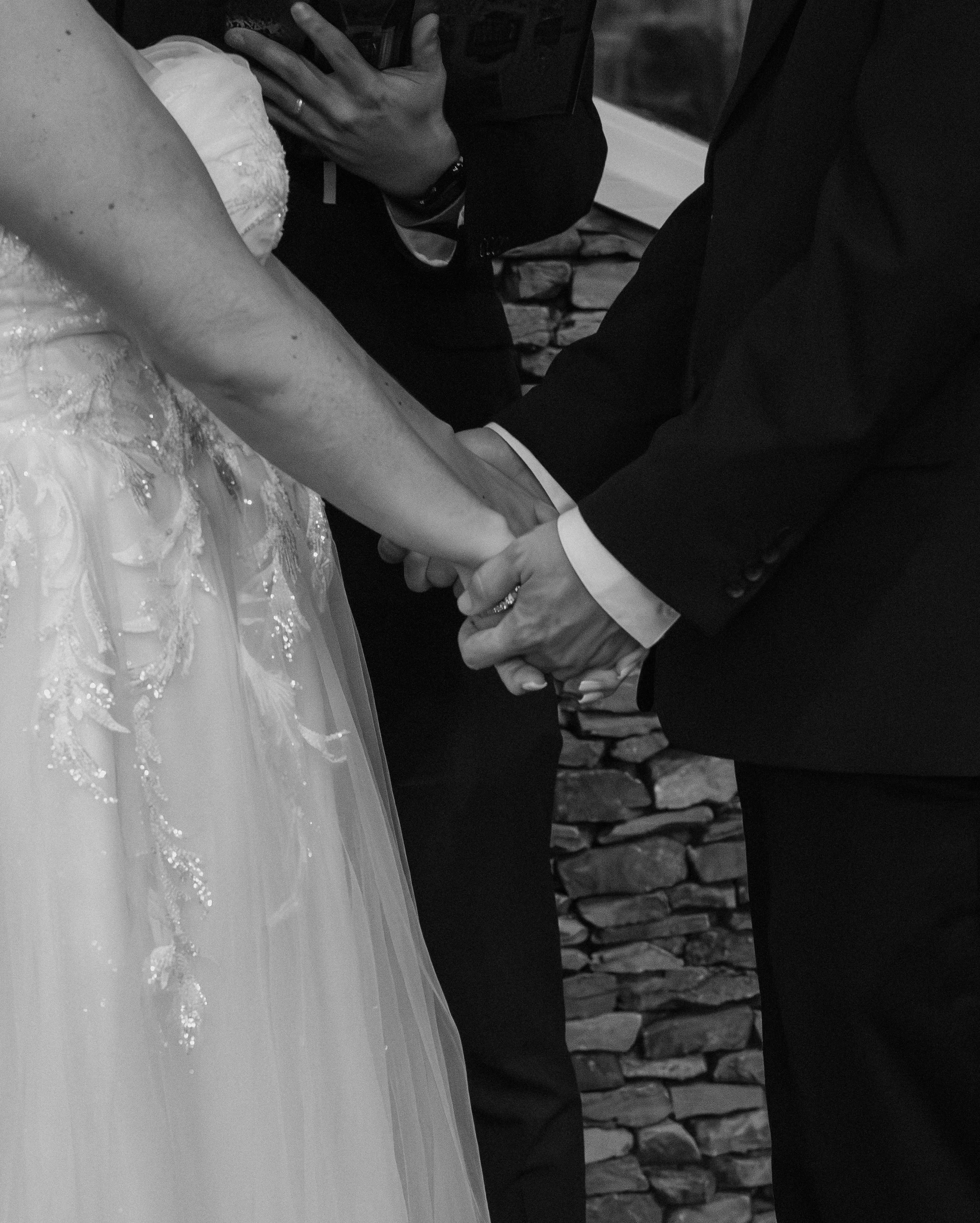 A bride and groom are holding hands during their wedding ceremony. The bride is wearing a wedding gown with intricate embroidery, and the groom is dressed in a dark suit. The focus is on their hands as they exchange vows or rings. The setting appears