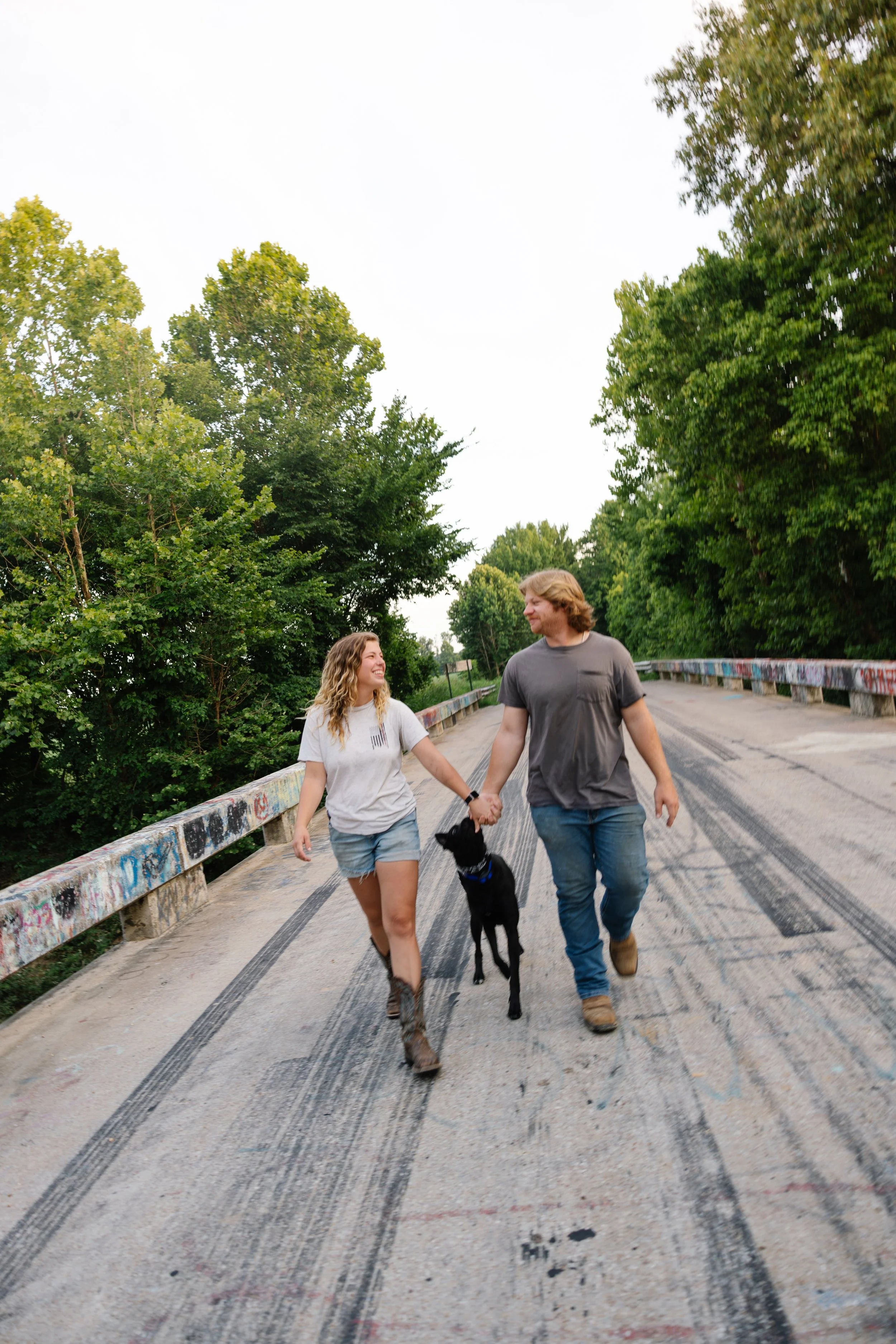 A young couple walks across a bridge holding hands, with a black dog between them. The woman has curly blonde hair and is wearing a white t-shirt and denim shorts. The man has shoulder-length hair and is wearing a gray t-shirt and jeans. The bridge h
