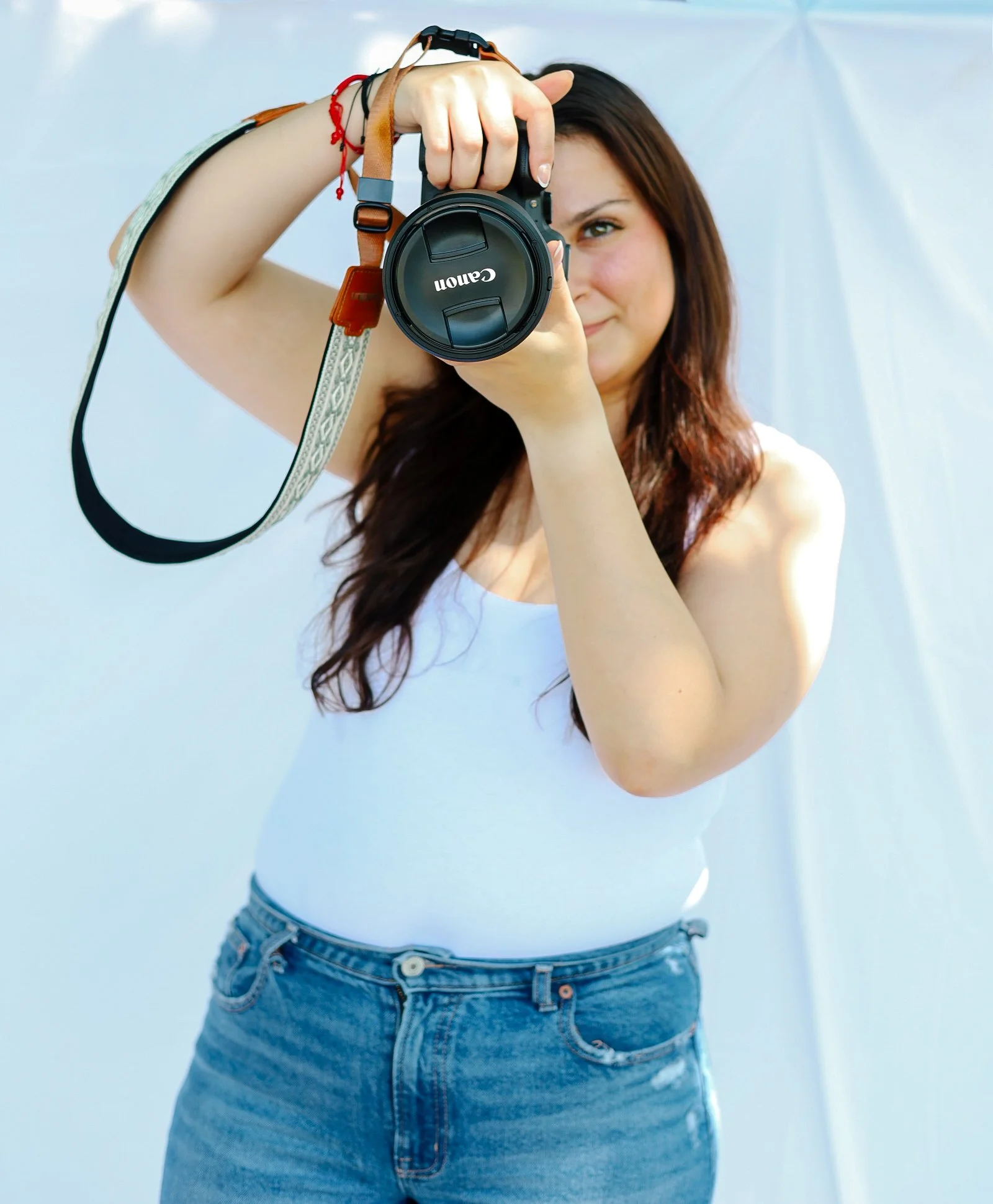 A woman with long brown hair wearing a white top and blue jeans, holding a DSLR camera aimed at the viewer, in front of a white background.