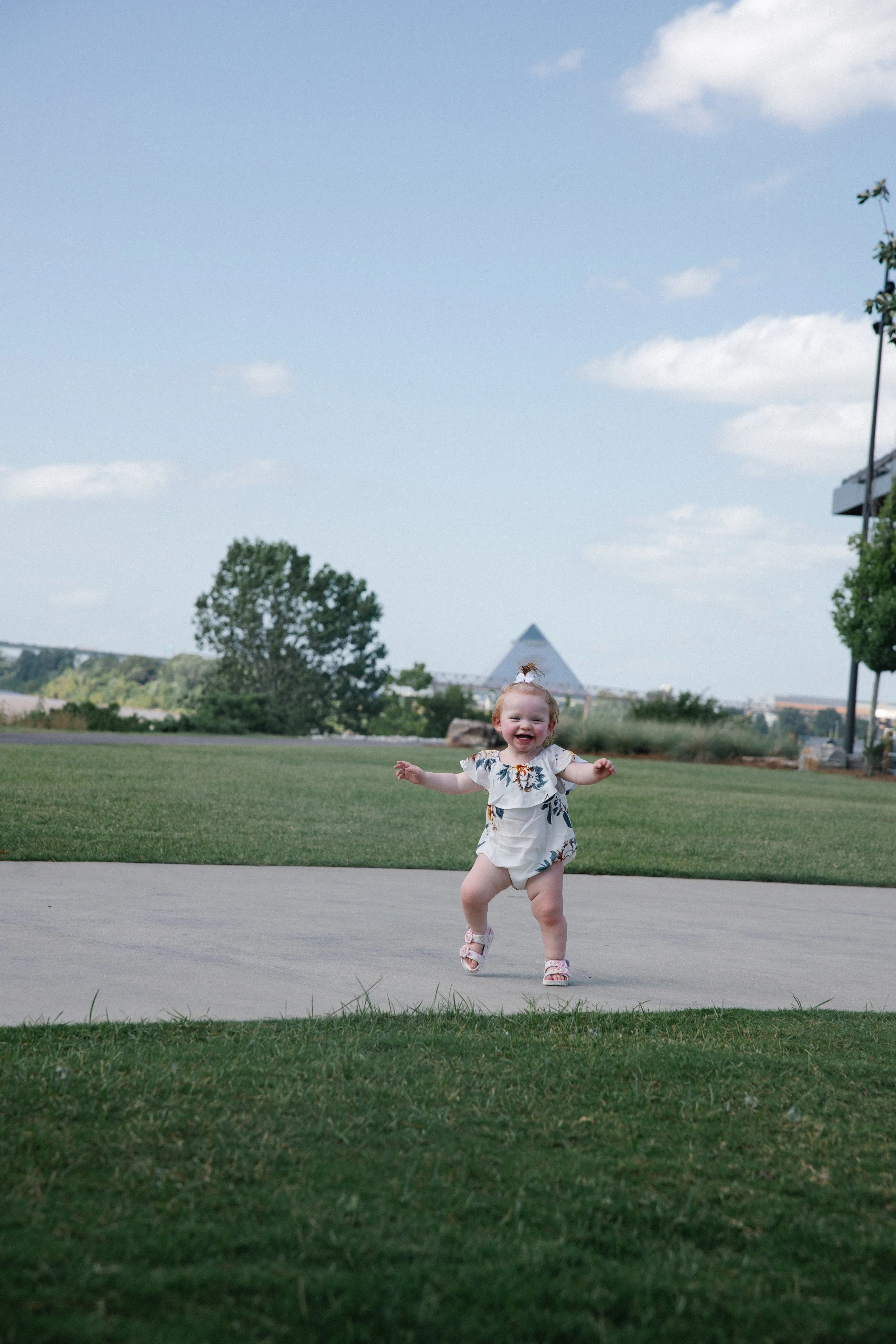 A smiling toddler girl running on a paved pathway in an outdoor park area, with trees, a grassy field, and a large pyramid-like structure in the background. The sky is mostly clear with some clouds.