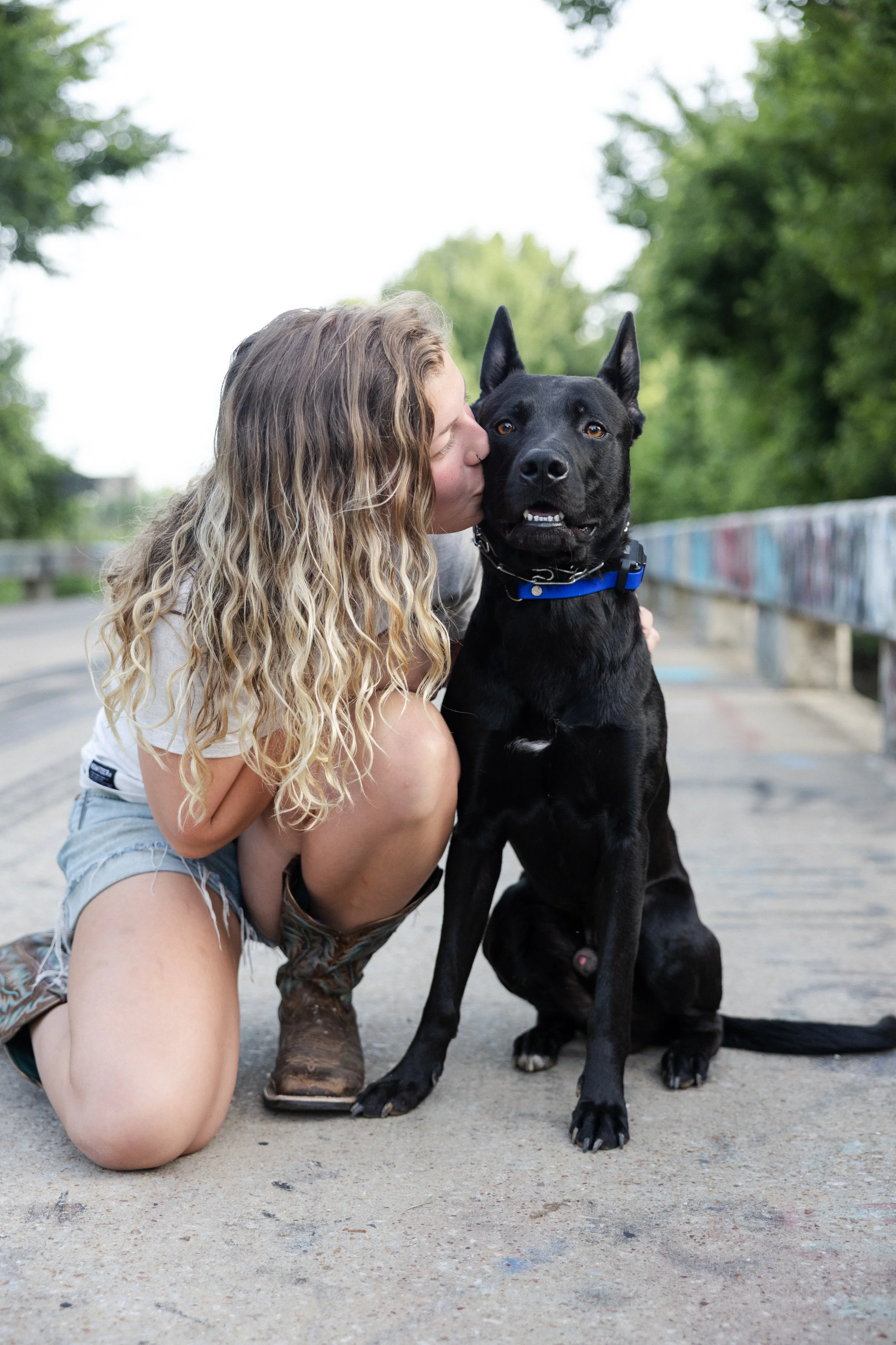 A young woman with long wavy blonde hair kneeling and kissing a large black dog with pointed ears on the head. The woman is wearing a white t-shirt and shorts, and the dog has a collar and leash. They are outdoors on a paved area with graffiti-covere