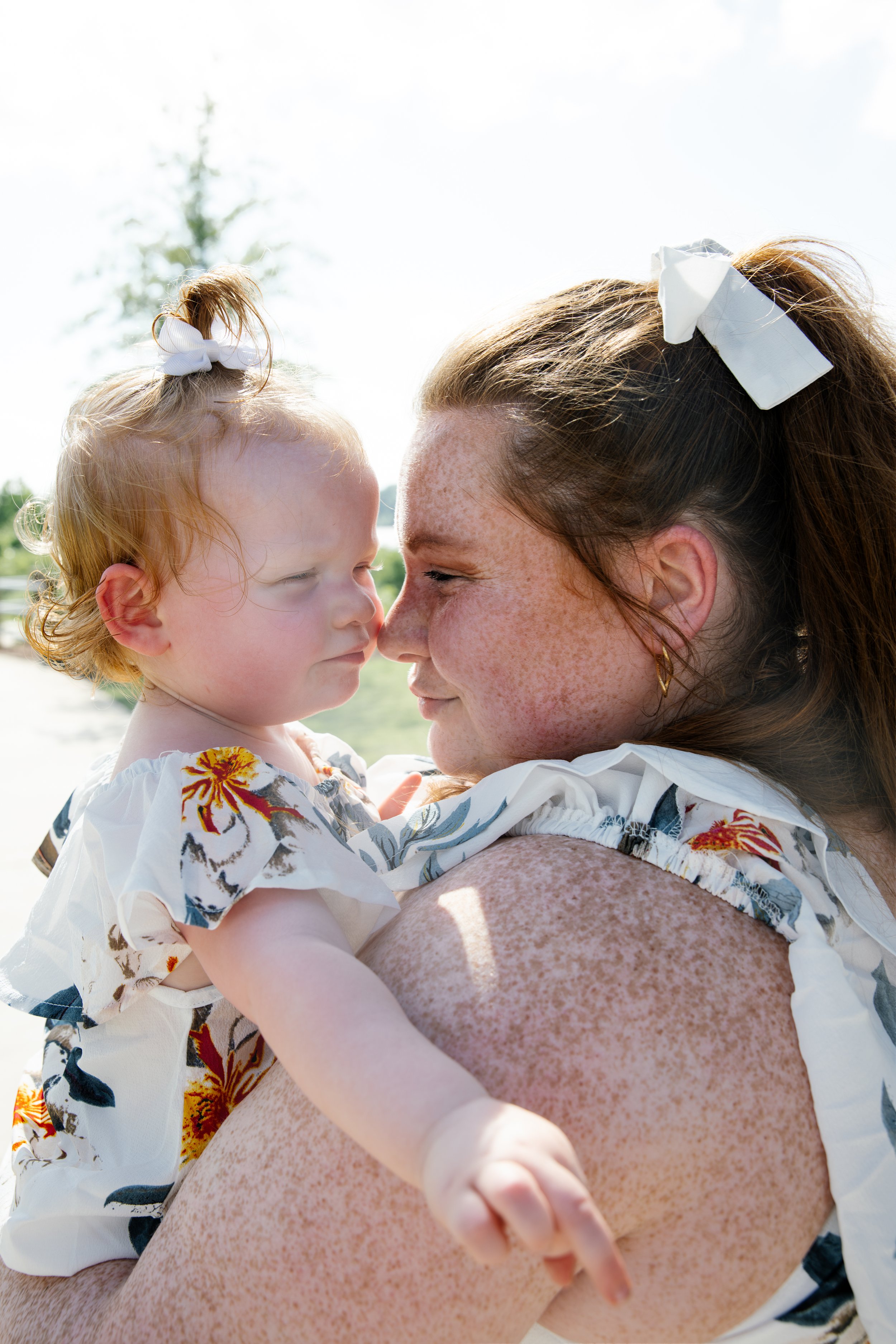A woman with freckles holds a young girl with a white bow in her hair close to her face outdoors on a sunny day. They are touching foreheads and smiling gently.