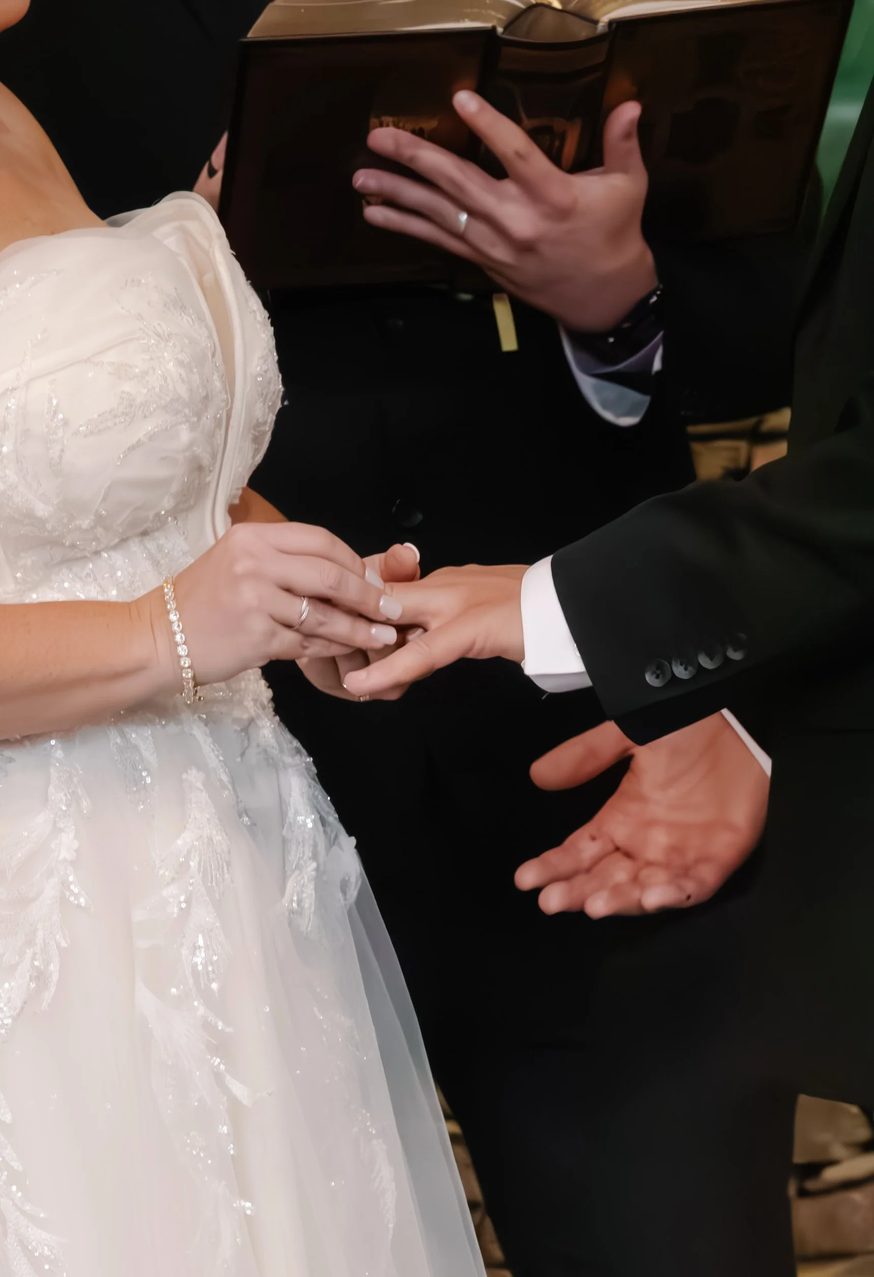 A bride and groom holding hands during a wedding ceremony. The bride wears a white dress with lace details, a bracelet, and has a ring on her finger. The groom is dressed in a black suit with a white shirt and a black tie, and is holding the bride's 