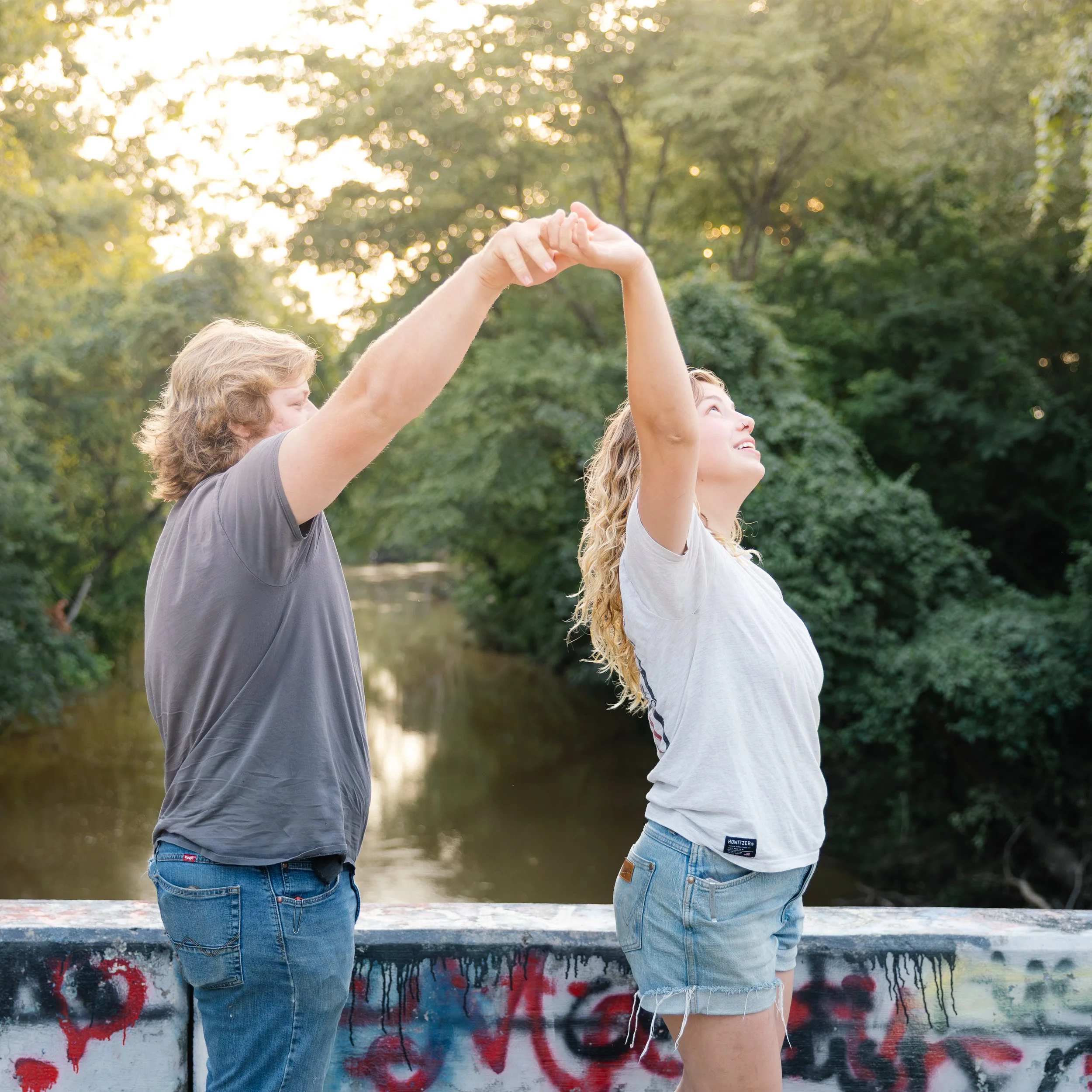 Two women standing outdoors at sunset, holding hands raised together, smiling, with trees in the background.